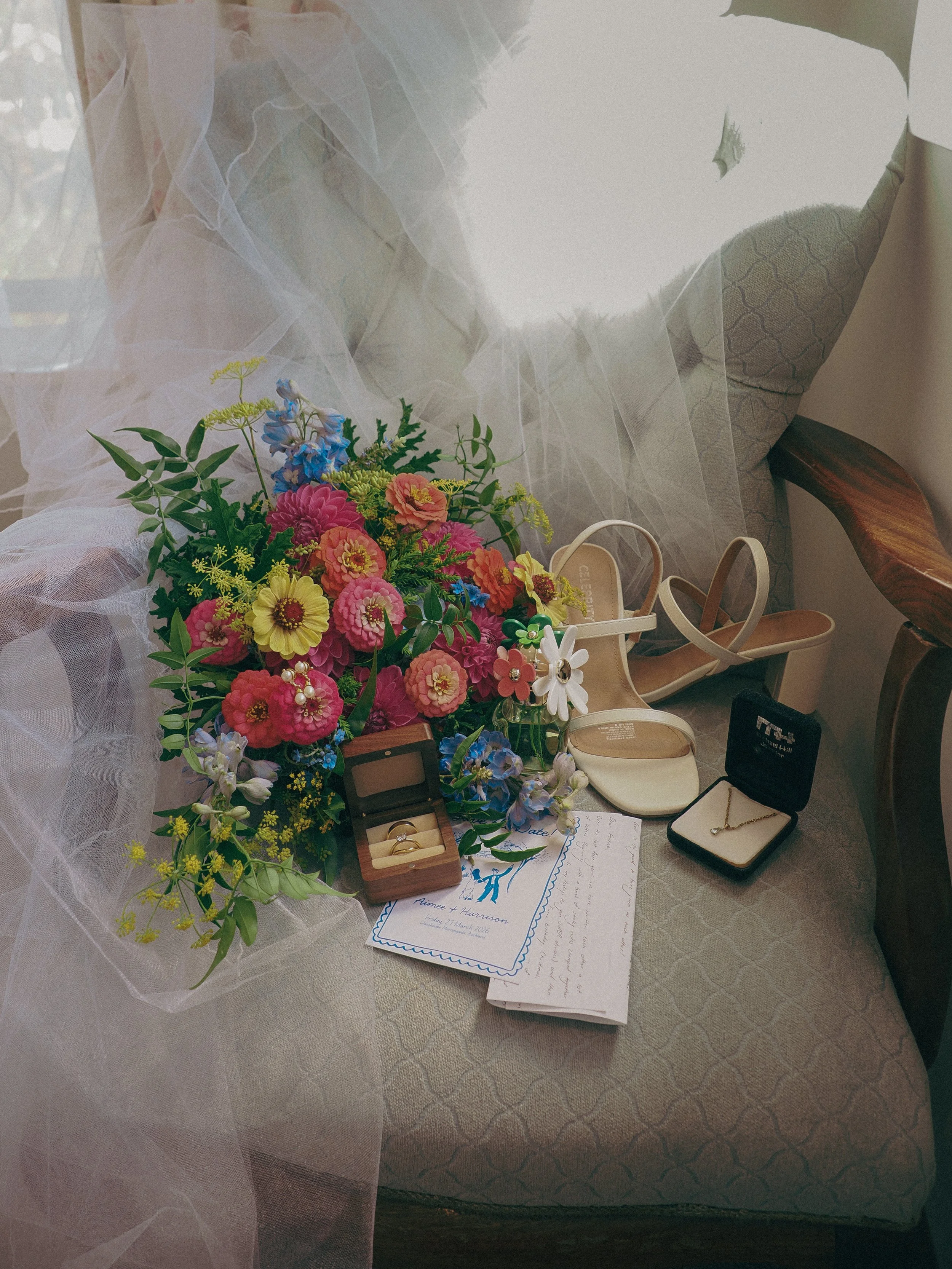 Wedding items arranged on a table including a bouquet of colorful flowers, a wedding invitation, a ring box with rings, shoes, and jewelry.