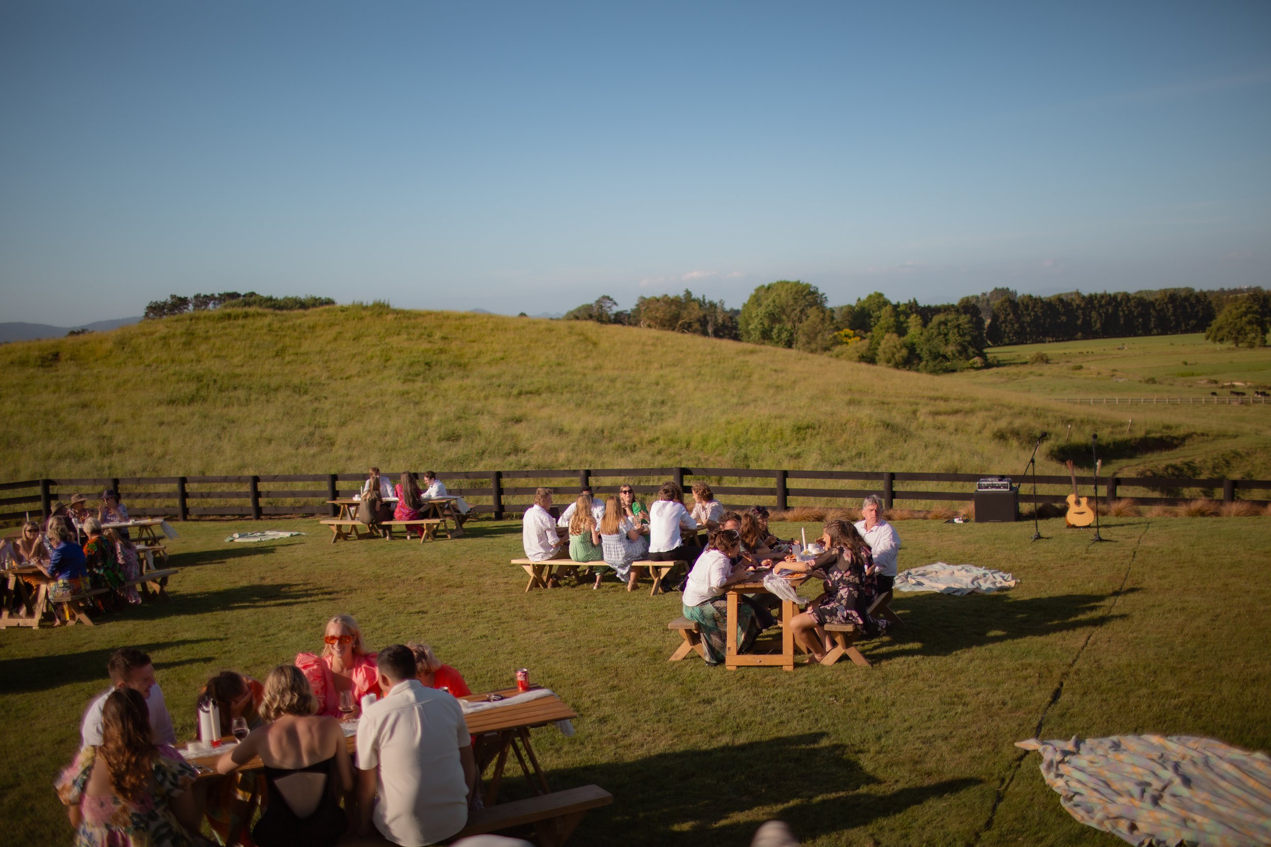 People sitting at picnic tables outdoors in a grassy field during late afternoon or early evening, with musical instruments set up nearby, and rolling green hills in the background.