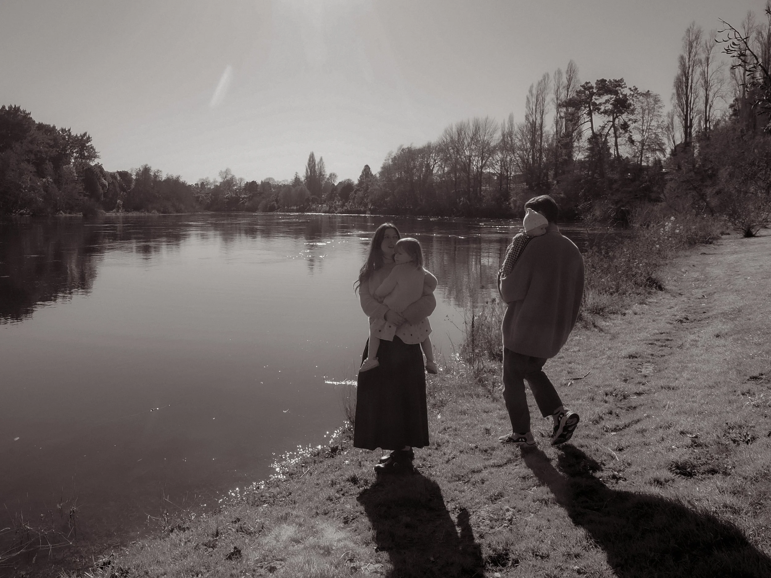 A black and white photograph of three people by a river. A woman is holding a child, and a man is walking nearby, carrying another child. Trees line the background along the riverbank. Family Photography Waikato New Zealand. Family Photography 