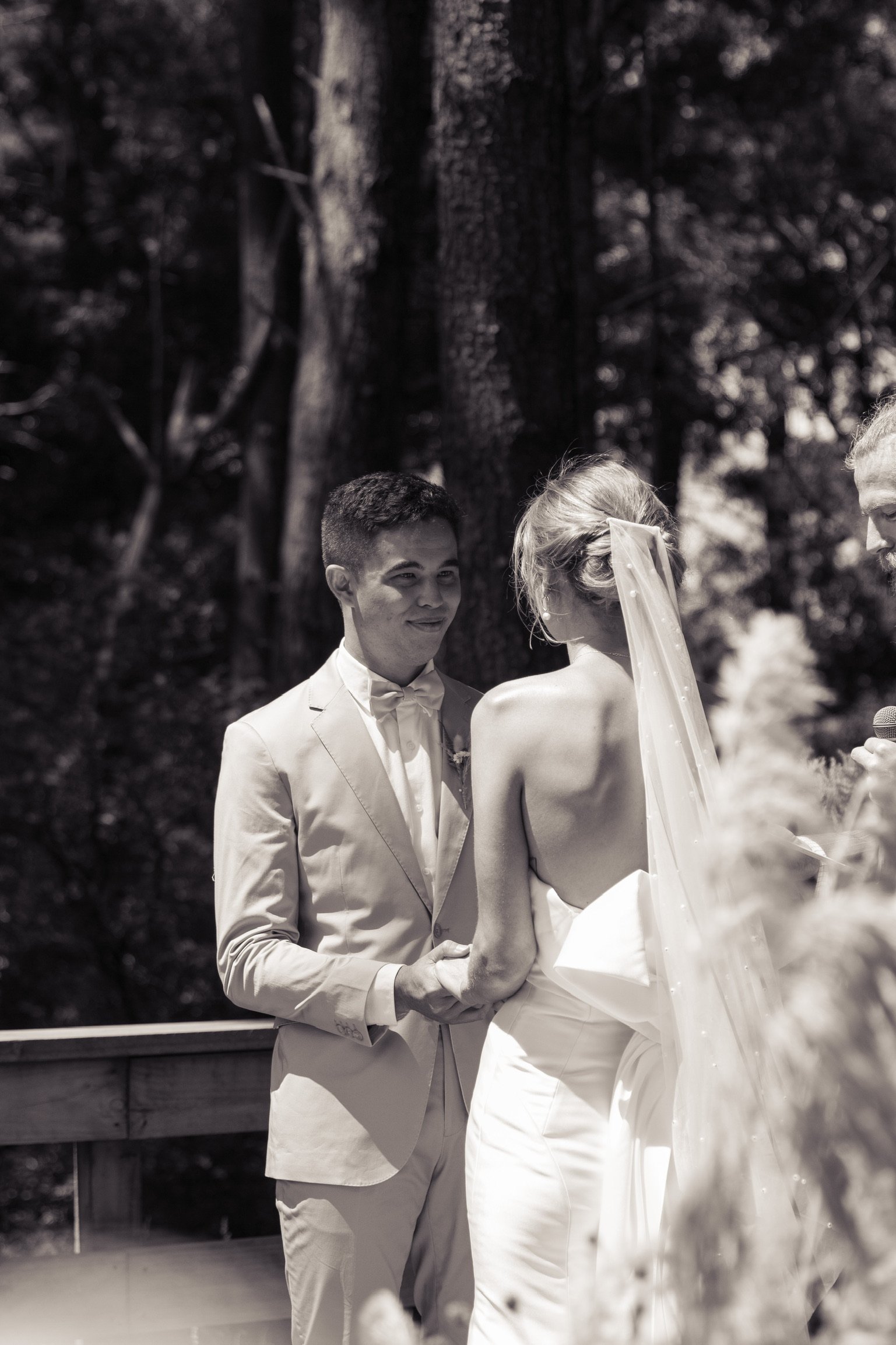 A black and white photo of a wedding ceremony, showing a groom holding hands with a bride who is wearing a veil and strapless dress, outdoors surrounded by trees.  Wedding Photography Waikato New Zealand. Wedding Photography Hamilton New Zealand. 