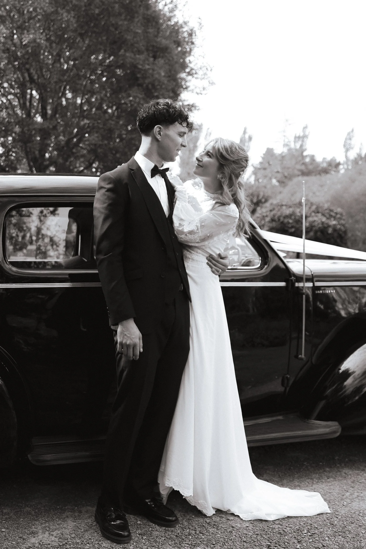 A black-and-white photograph of a bride and groom standing close near a vintage car. The groom is dressed in a tuxedo and the bride in a long wedding dress, looking at each other with joy, outdoors with trees in the background. Wedding Photography 
