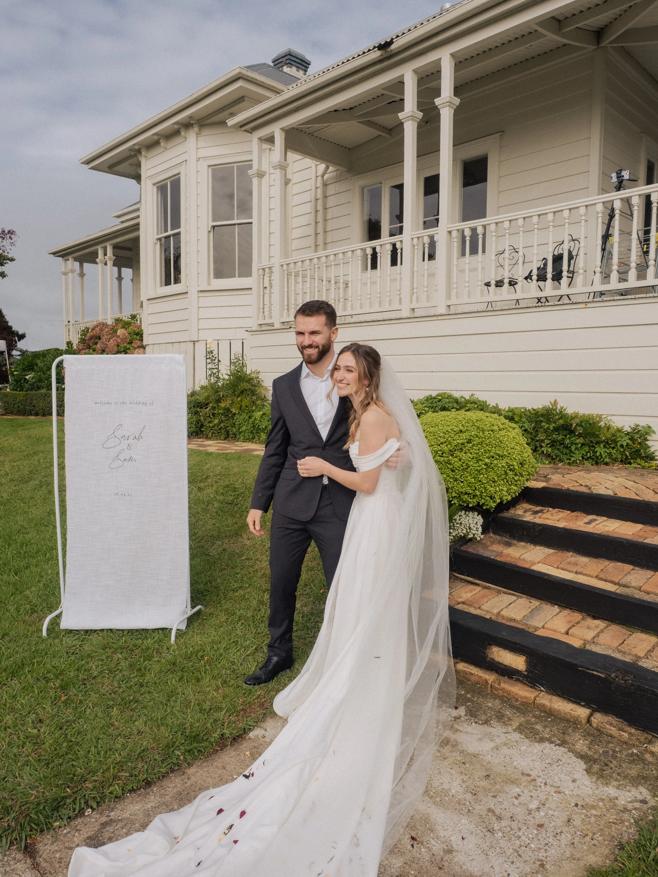 A newlywed couple, a man in a black suit and a woman in a white wedding dress, walking outside in front of a white house with a porch, smiling and holding each other.  Wedding Photography Waikato New Zealand. Wedding Photography Hamilton New Zealand.