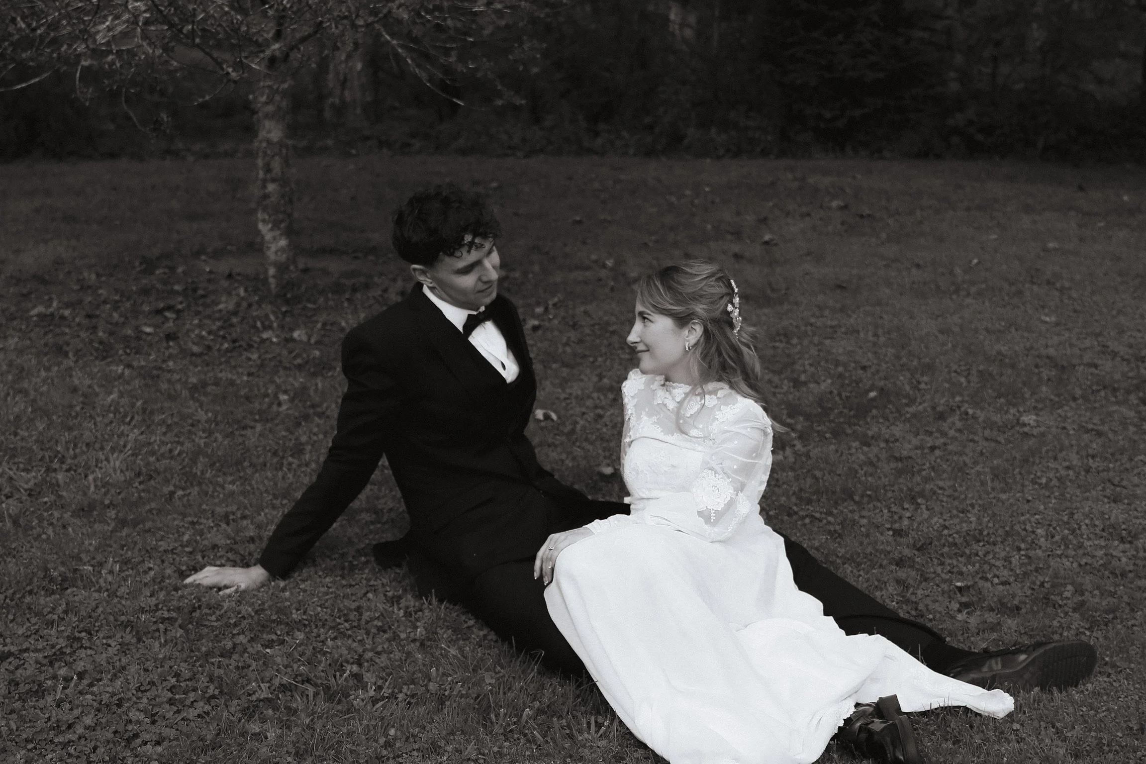 A couple dressed in wedding attire sitting on the ground outdoors at night, with trees in the background. The groom is wearing a tuxedo and the bride is in a white wedding dress, and they are looking at each other.