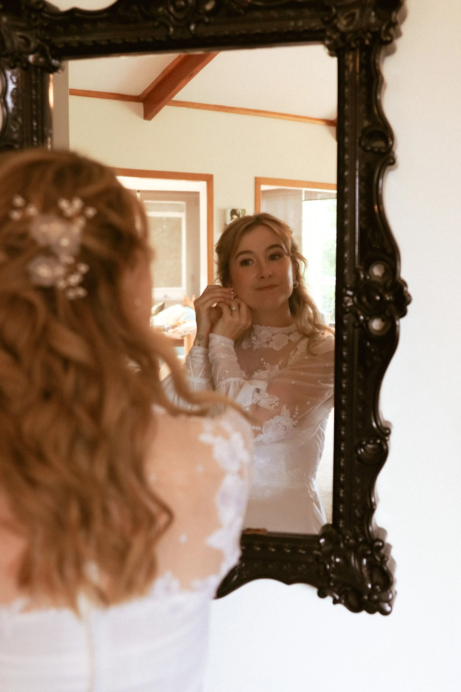 A woman in a white lace dress is seen looking into a mirror while adjusting her earring. Wedding Photography Waikato New Zealand. Wedding Photography Hamilton New Zealand