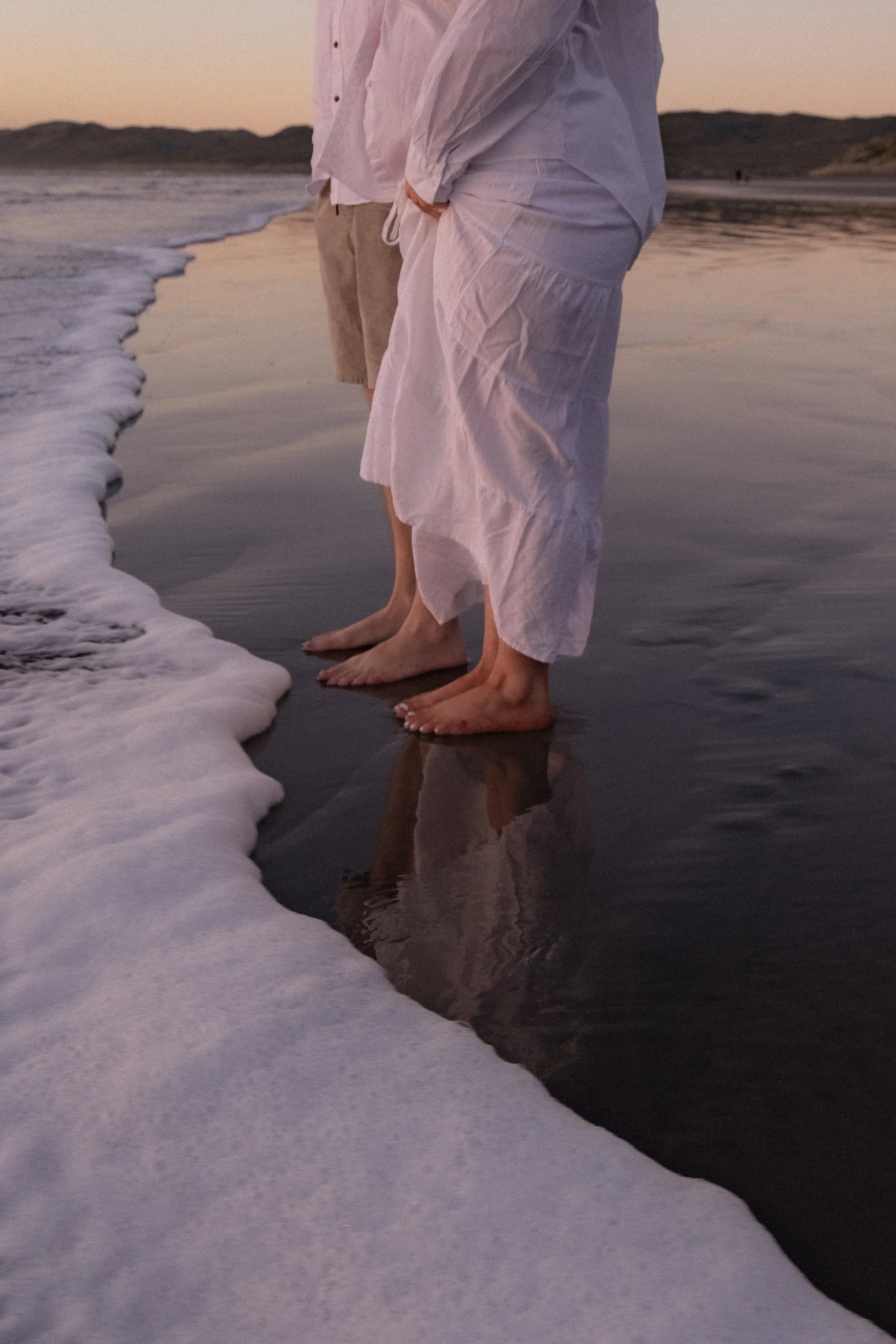 Two people standing barefoot at the edge of the ocean during sunset, with gentle waves washing over their feet and reflections visible on wet sand. Maternity Photography
