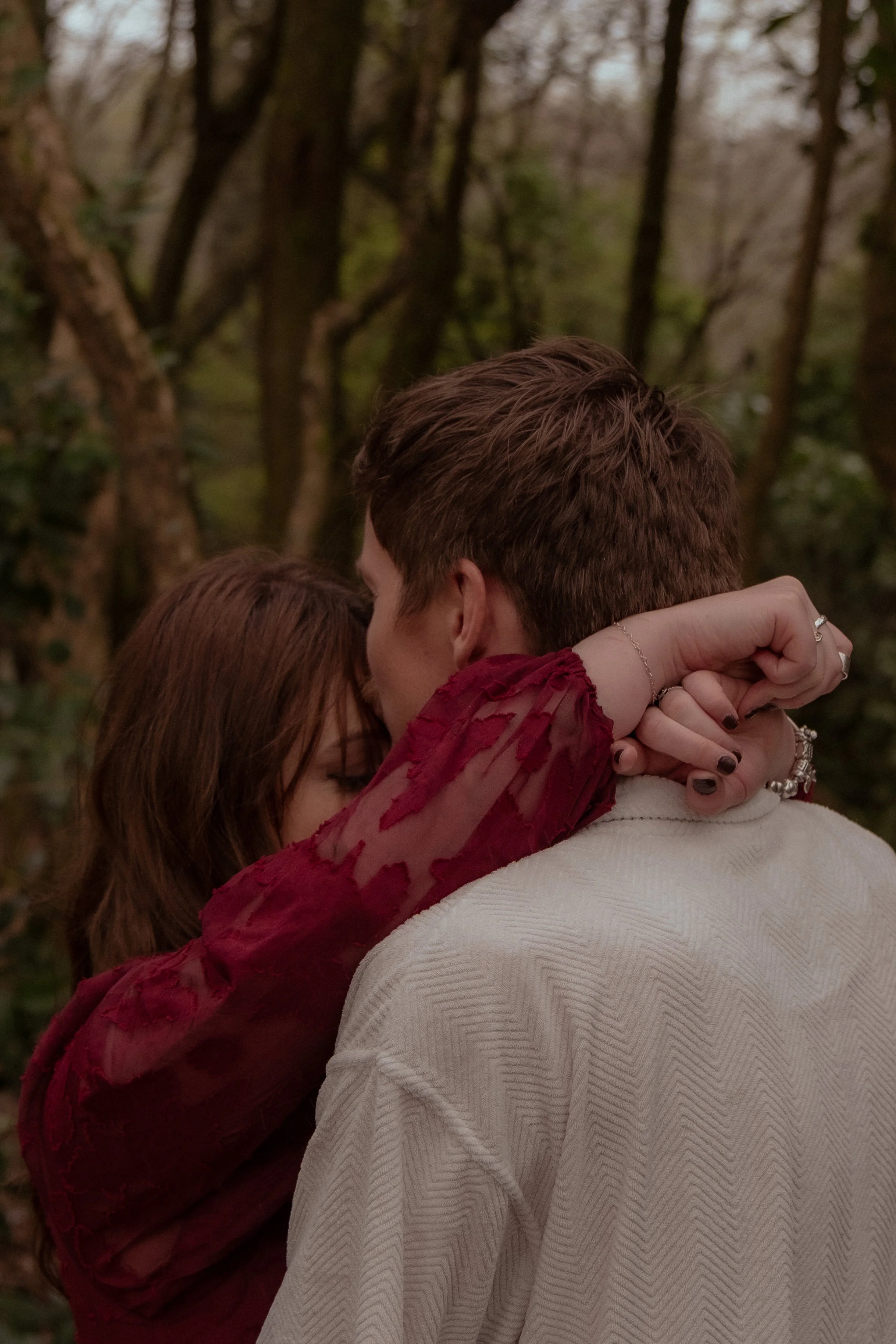 Close-up of a couple sharing an intimate moment outdoors, with a woman in a red, sheer, embroidered blouse and a man in a white shirt, surrounded by trees. Engagement photography Hamilton New Zealand. Whangarei Photographer
