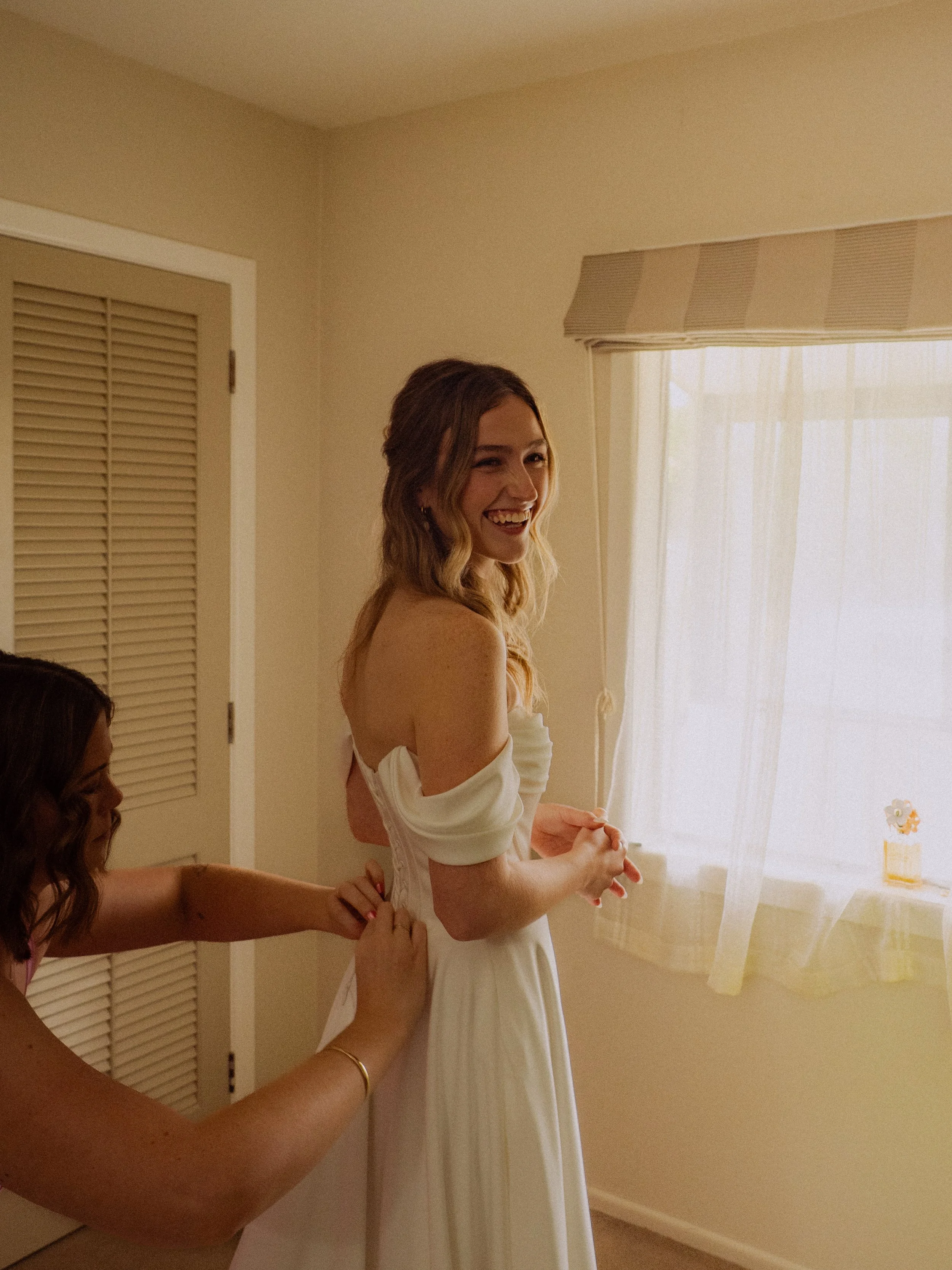 A woman in a white dress smiling happily as someone helps her with her dress inside a room with a window and cream-colored walls.  Wedding Photography Waikato New Zealand. Wedding Photography Hamilton New Zealand. Northland Wedding Photography