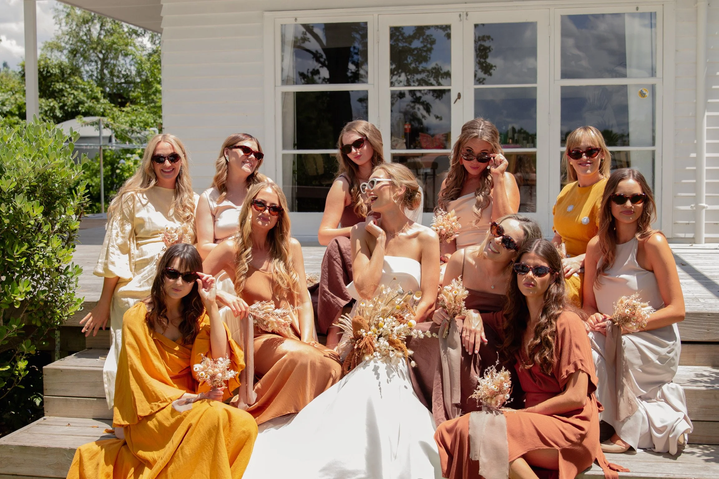 A group of women dressed in colorful dresses sitting and standing on wooden steps outside a white house with large windows, holding bouquets, posing and smiling, with some wearing sunglasses.