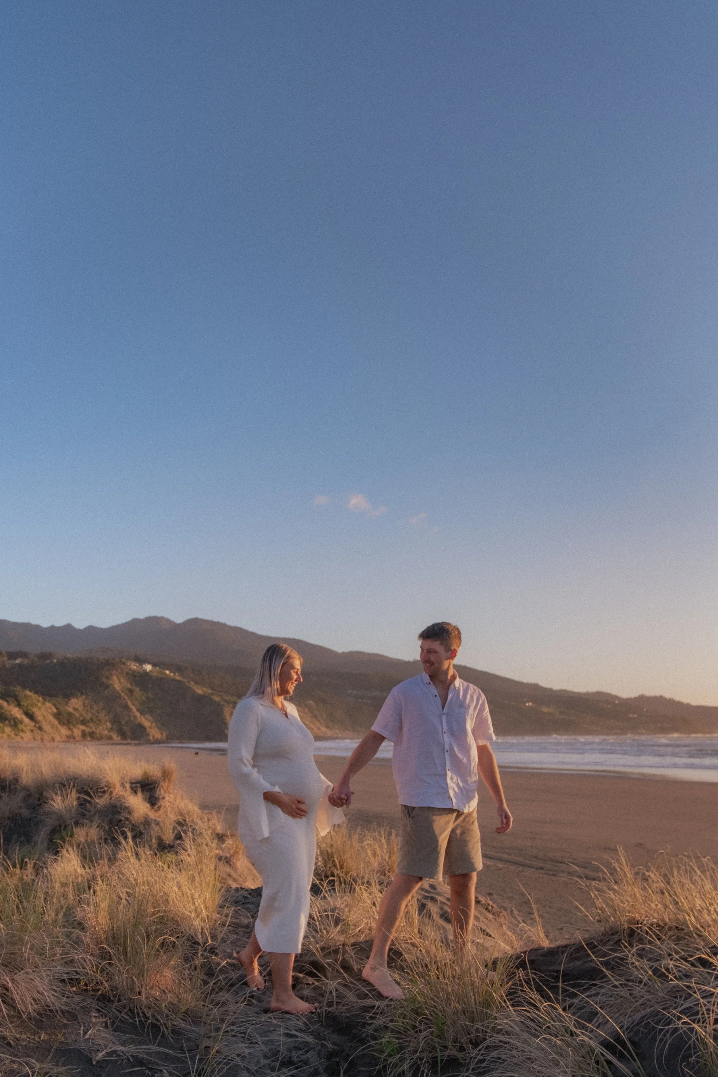 A pregnant woman and a man walking on a beach holding hands during sunset, with dunes and mountains in the background. Maternity Shoot. Raglan New Zealand