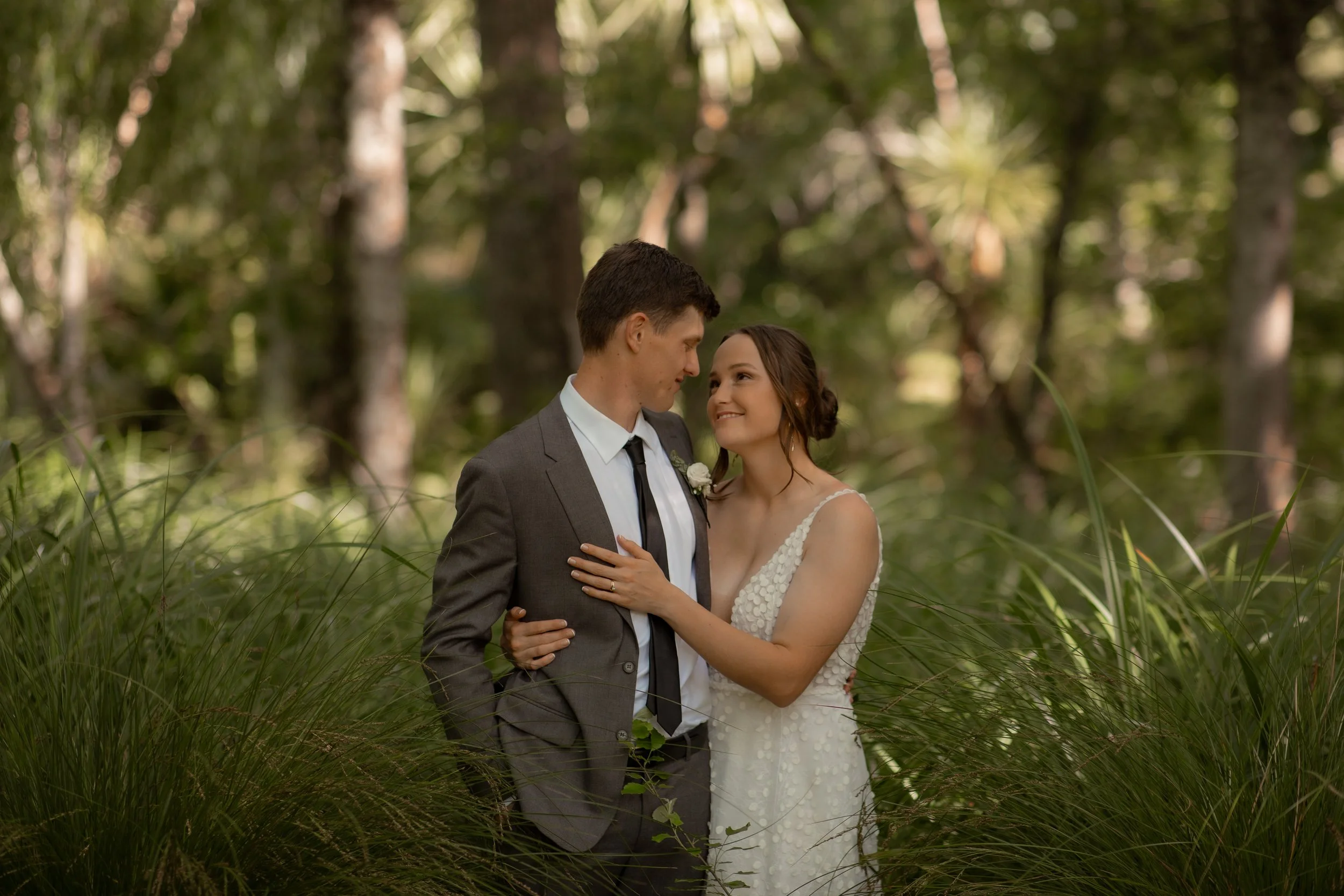 A bride and groom standing closely in a forested area, sharing an intimate moment and smiling at each other.