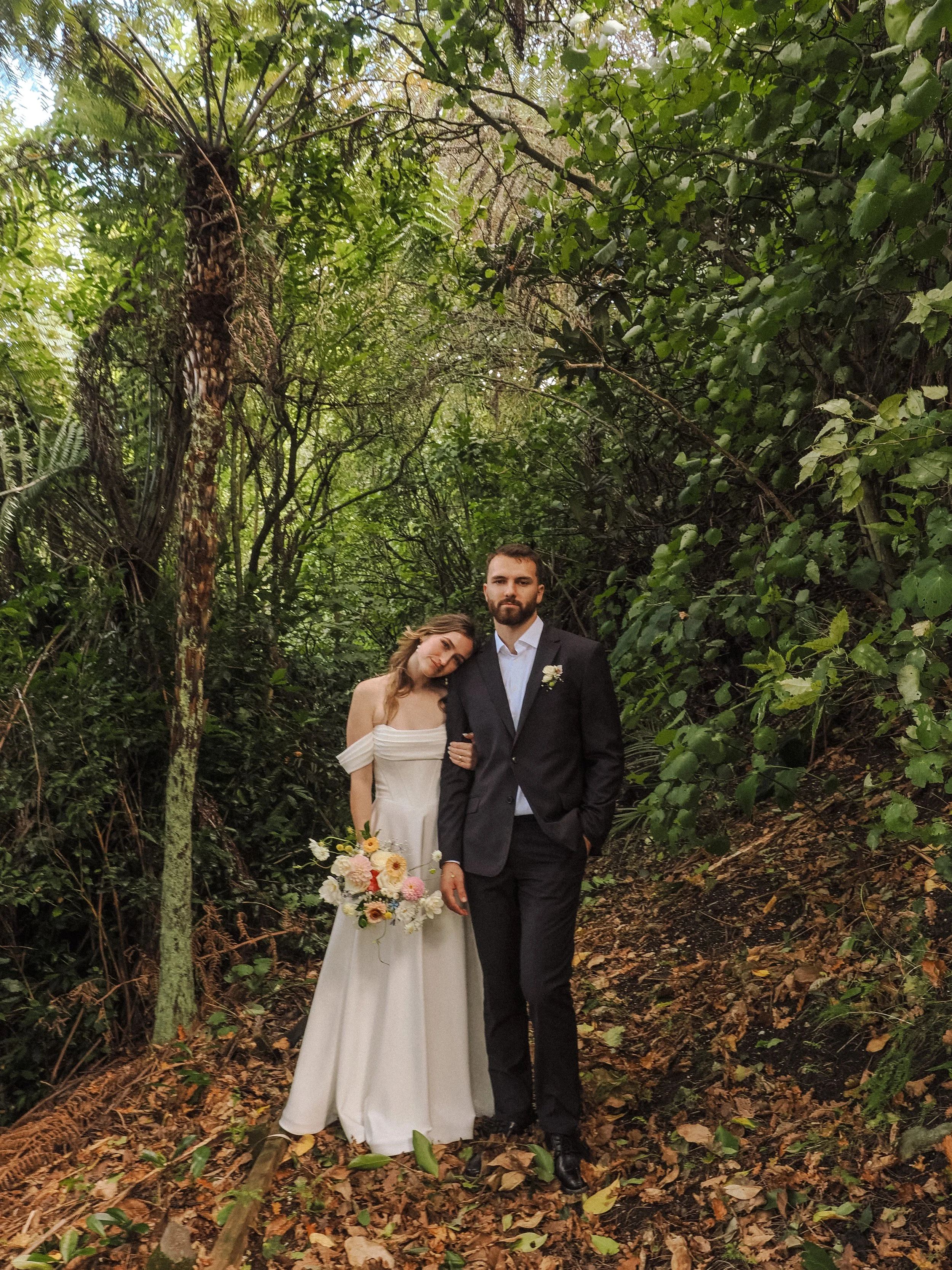 A bride and groom standing together in a lush, green forest, with the bride holding a bouquet of flowers and leaning her head on the groom's shoulder.  Wedding Photography Waikato New Zealand. Wedding Photography Hamilton New Zealand. Northland 