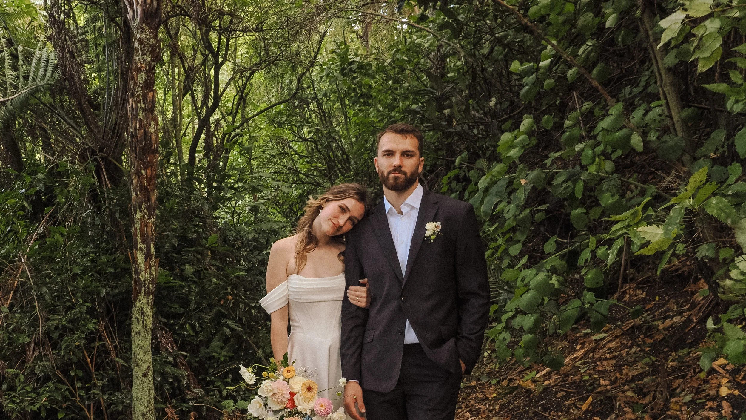 A bride and groom standing closely together in a lush, green forest. The bride, with brown hair, is leaning her head on the groom's shoulder and is holding a bouquet of white and pink flowers. The groom, with a beard, is wearing a black suit and whit