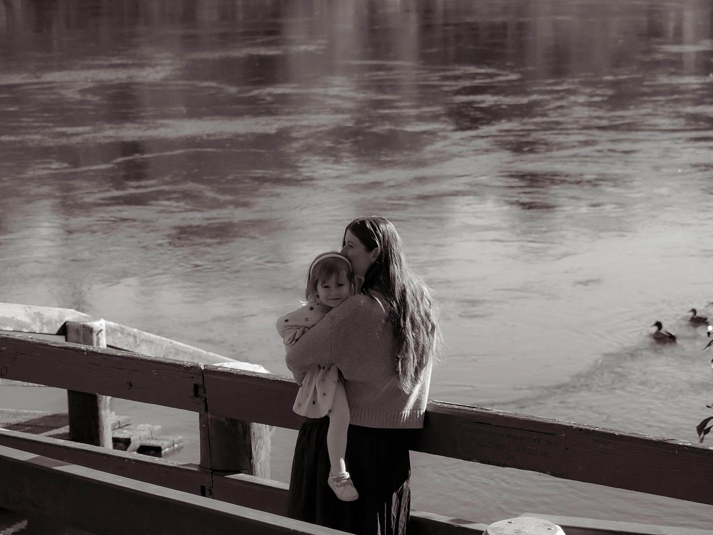 A woman holding a young girl by the water's edge, with ducks swimming in the water in the background.  Family Photography Waikato New Zealand. Family Photography Hamilton New Zealand. Northland Wedding Photography