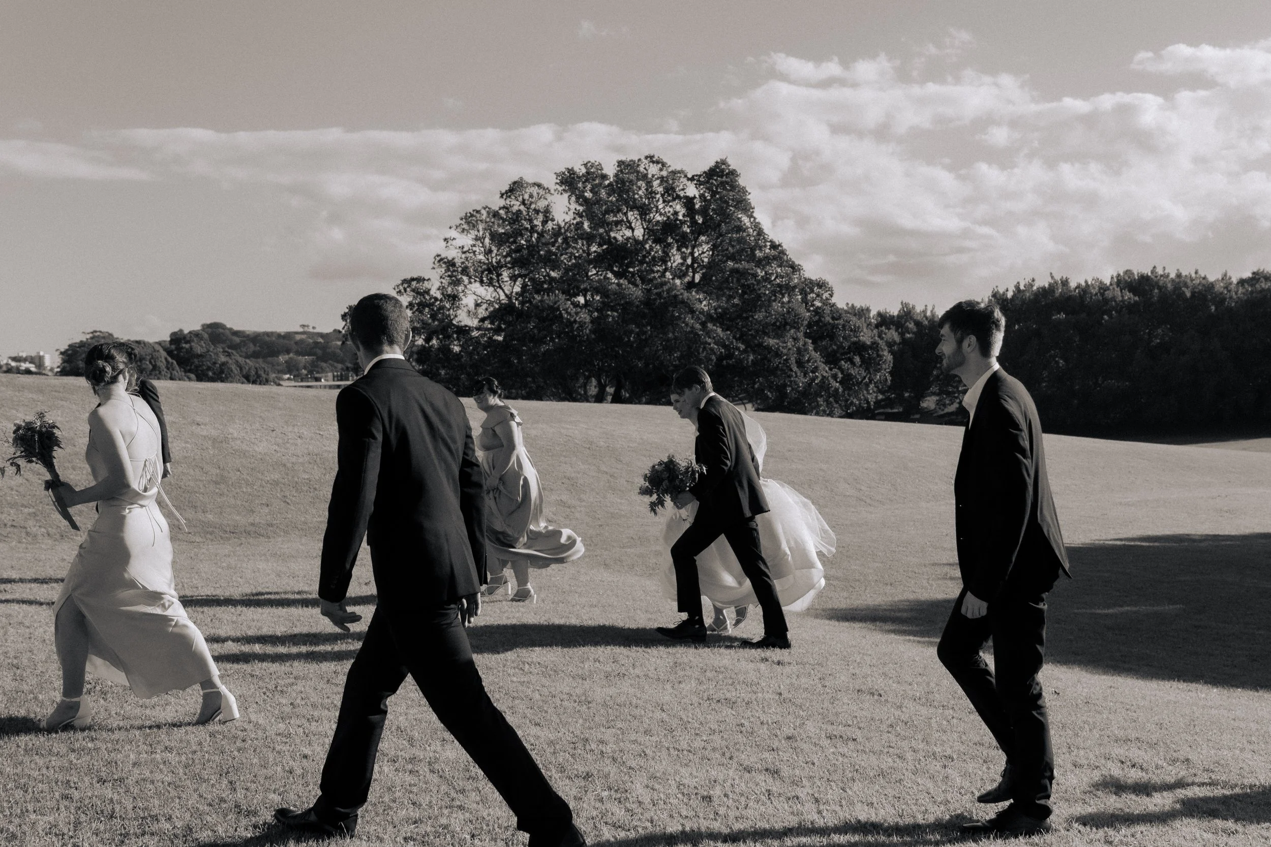 A black-and-white photo of a group of people walking across a grassy field, with trees and a partly cloudy sky in the background. Some are dressed in formal attire, suggesting a wedding or special event.