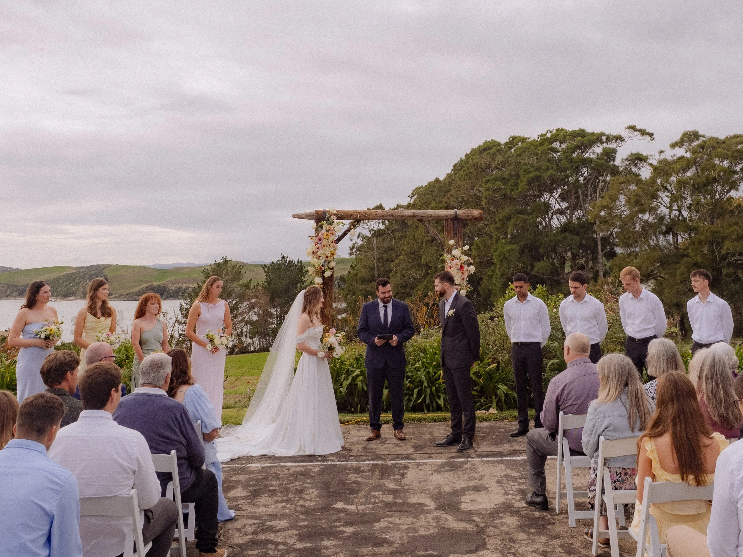 An outdoor wedding ceremony with a bride and groom facing each other under a wooden arch decorated with flowers, surrounded by bridesmaids and groomsmen, with seated guests watching near a body of water and trees in the background.