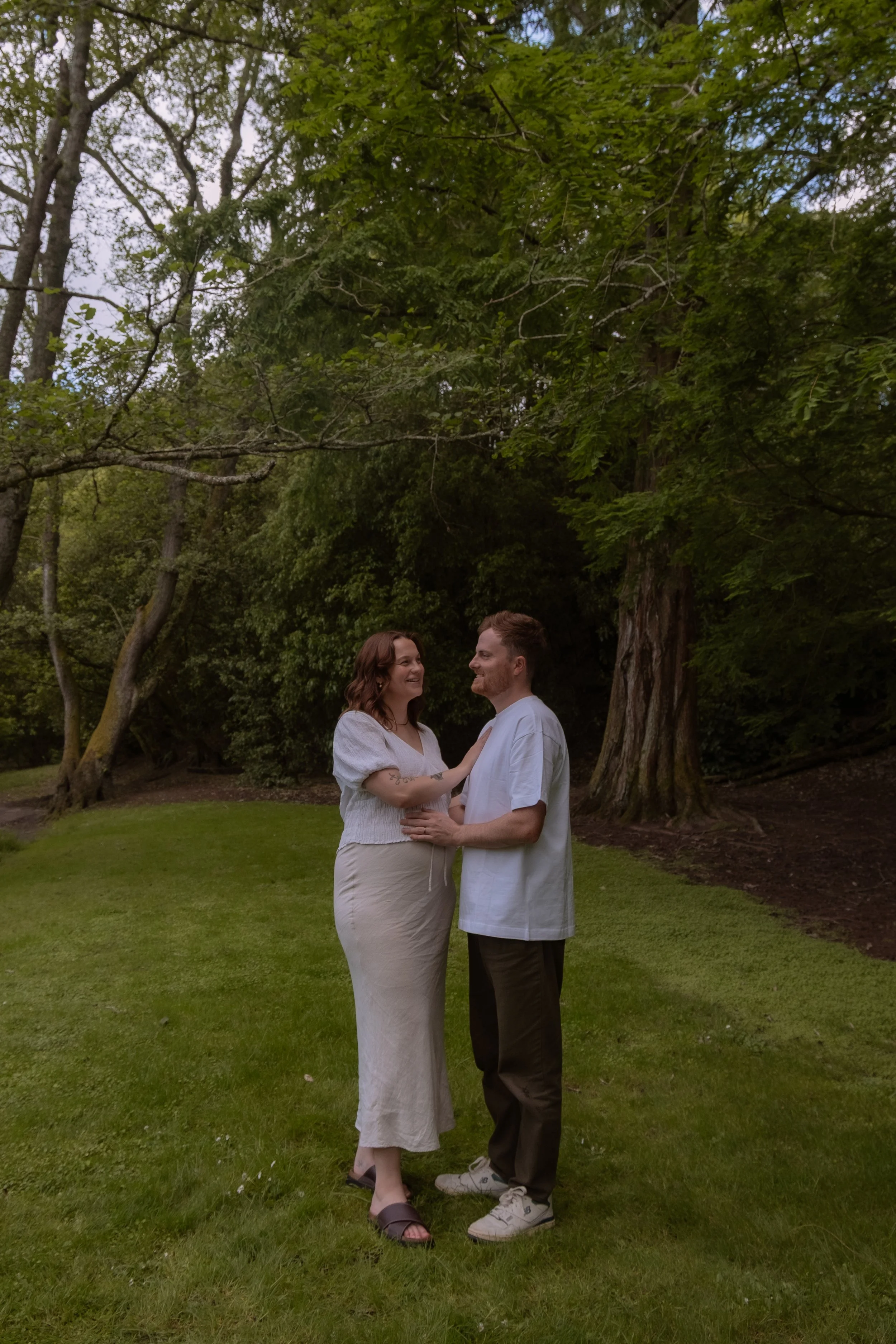 A couple standing on a grassy area, facing each other and smiling, surrounded by large trees in a park-like setting. Maternity Photography. Hamilton New Zealand Photographer