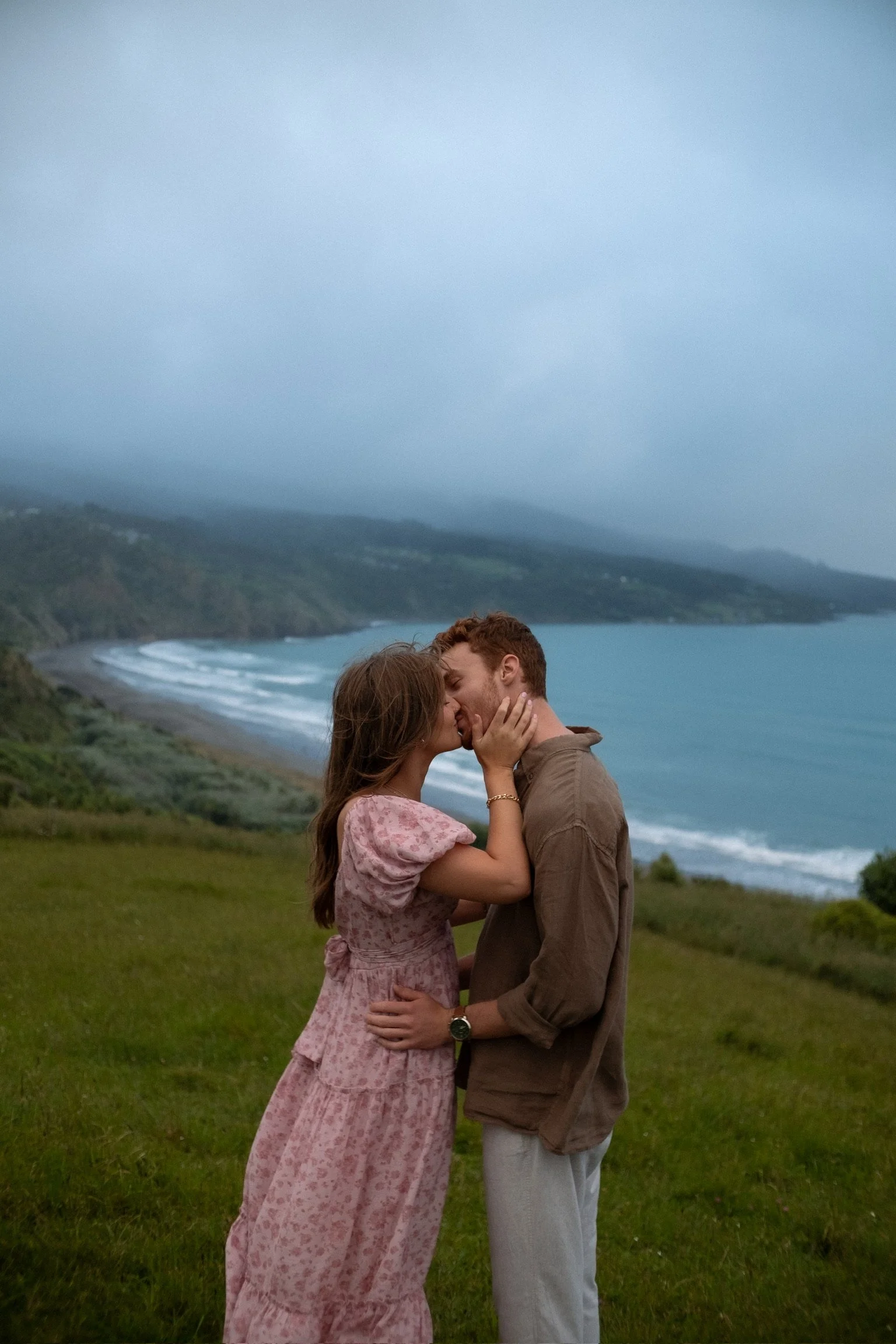 A couple sharing a kiss on a grassy hill overlooking the ocean and coastline under an overcast sky. Couples photography Raglan New Zealand. Engagement photography Hamilton New Zealand. Whangarei New Zealand Couples photographer