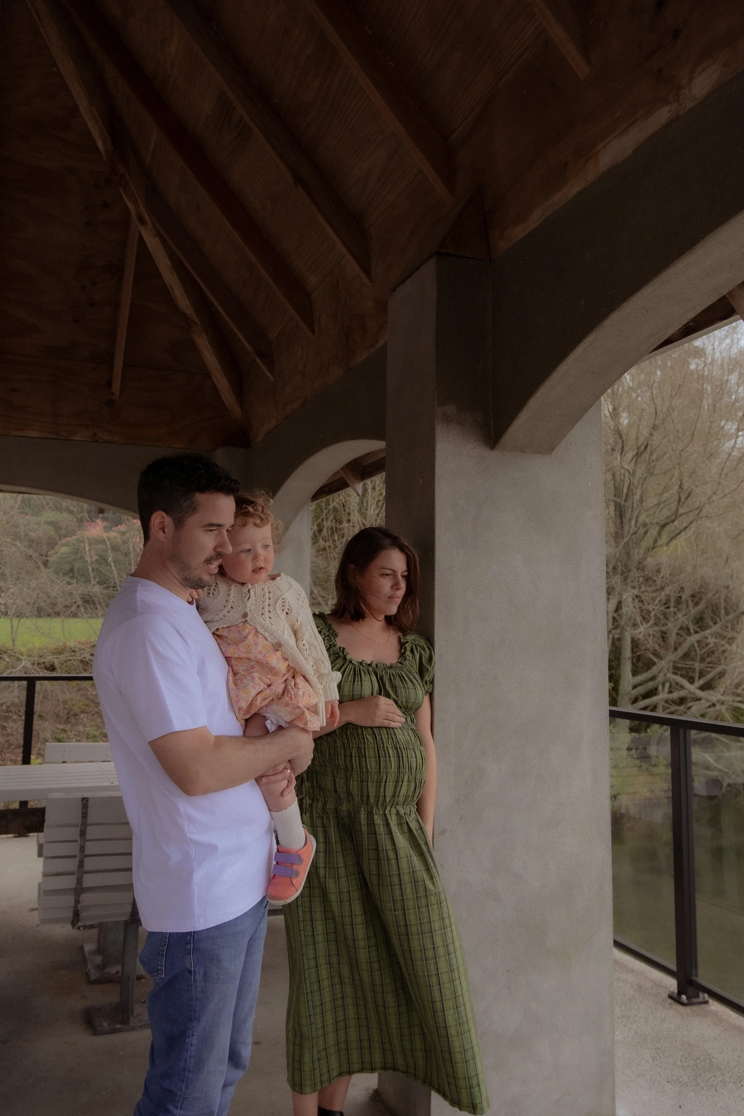 Family of three, a man, a woman, and a young girl, standing under a wooden pavilion near a river, with trees in the background. Family/Maternity photography Hamilton New Zealand