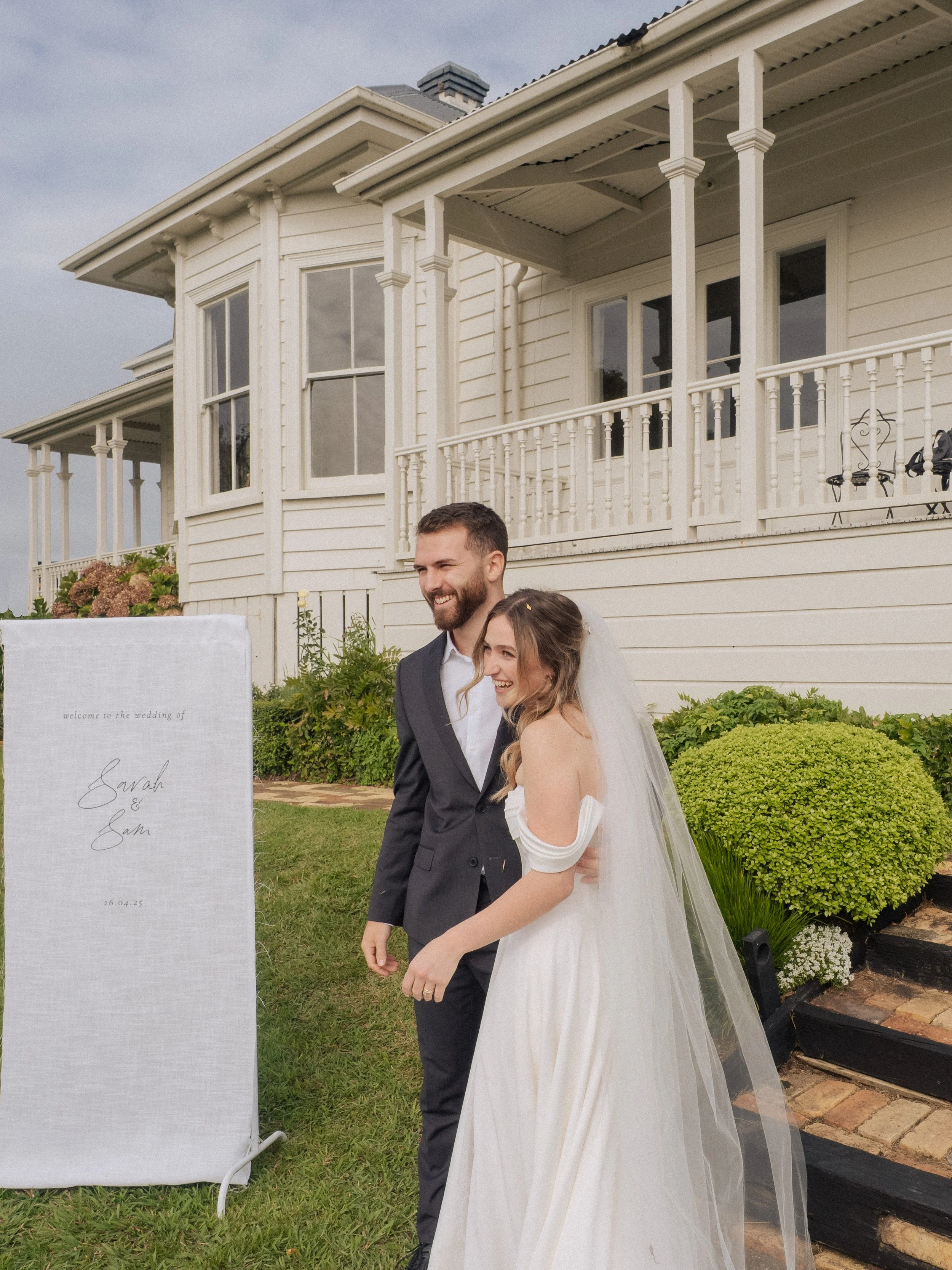 A bride and groom standing outside a white house, smiling at a wedding event.  Wedding Photography Waikato New Zealand. Wedding Photography Hamilton New Zealand. Northland Wedding Photography
