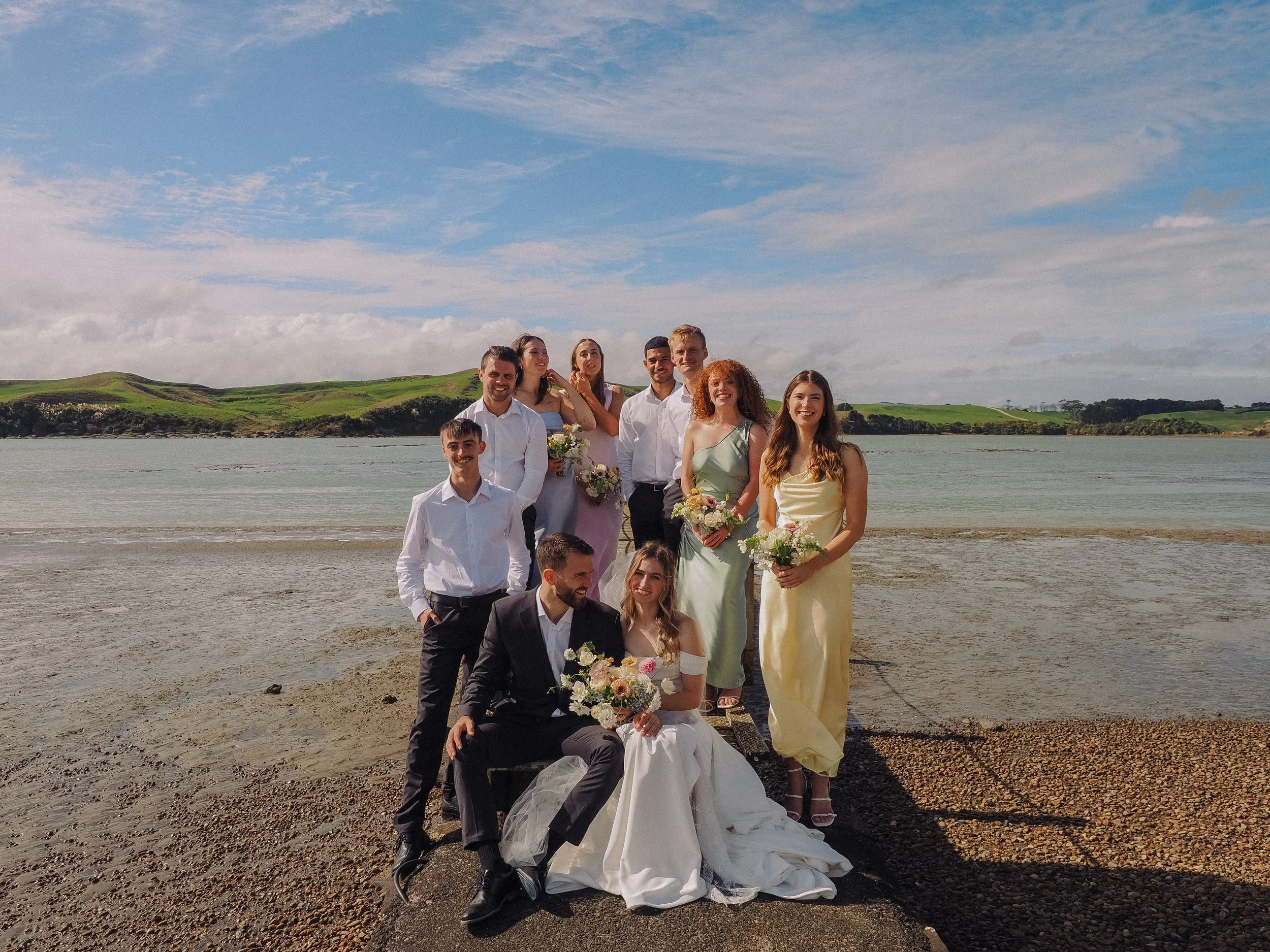A group of people dressed in wedding attire standing and sitting on a riverbank with water and green hills in the background, under a partly cloudy sky.  Wedding Photography Waikato New Zealand. Wedding Photography Hamilton New Zealand. Northland