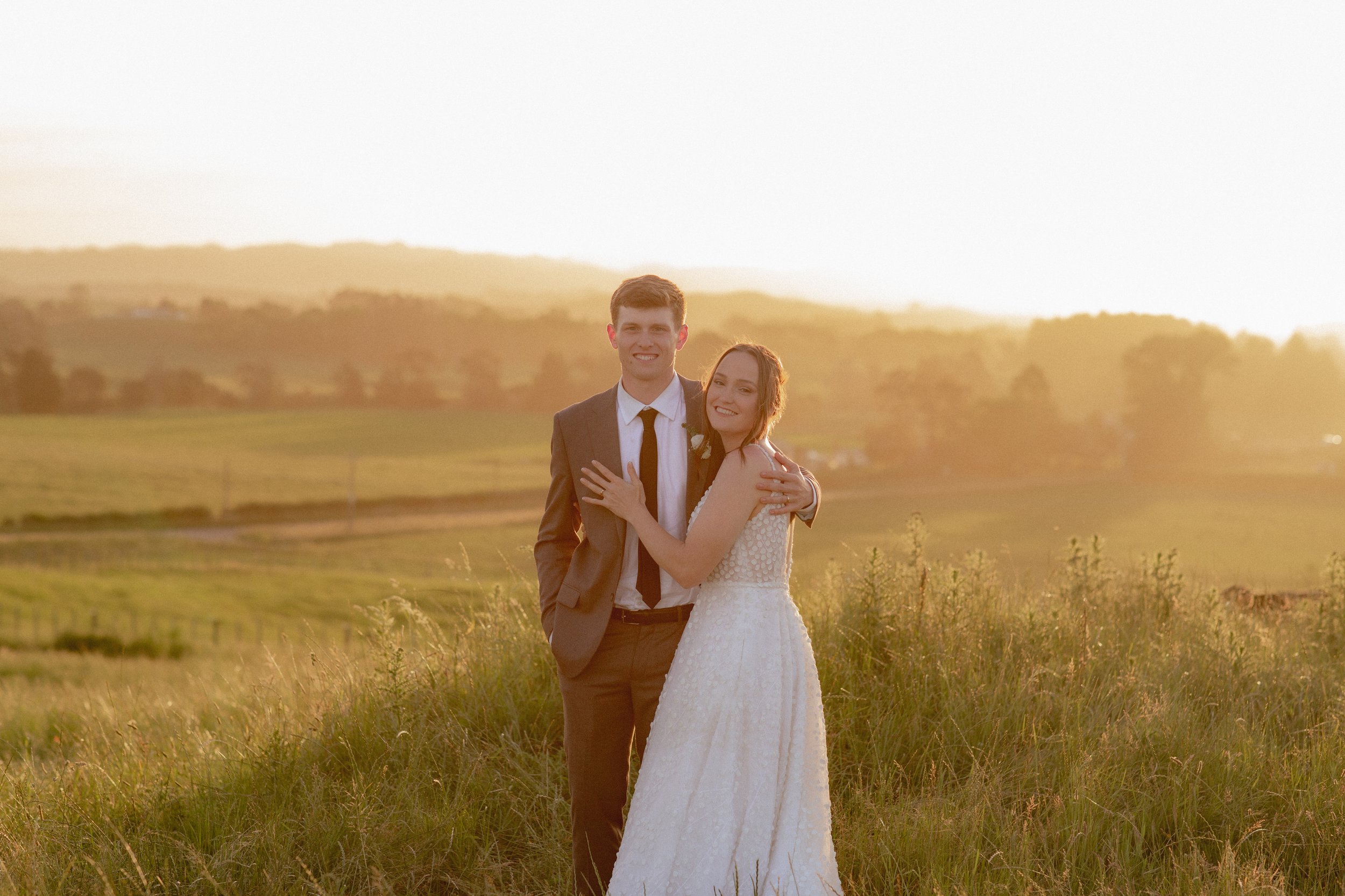 A young couple, dressed in wedding attire, standing in a grassy field during sunset with rolling hills in the background.  Wedding Photography Waikato New Zealand. Wedding Photography Hamilton New Zealand. Northland Wedding Photography