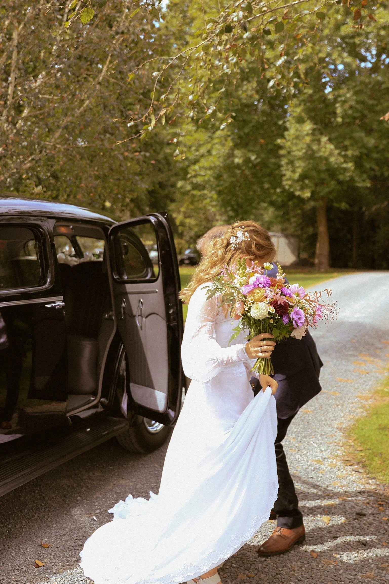 A bride in a white wedding dress holding a bouquet of flowers exits a black vintage car while a person helps her out. Wedding Photography Waikato New Zealand. Wedding Photography Hamilton New Zealand