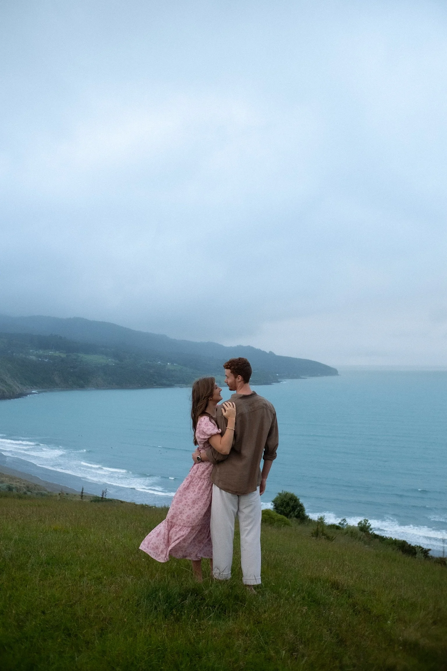 A couple standing on a grassy hill near the coast, embracing and smiling at each other, with a scenic view of the ocean, cliffs, and cloudy sky in the background. Couples photography Raglan New Zealand. Engagement photography Hamilton New Zealand.