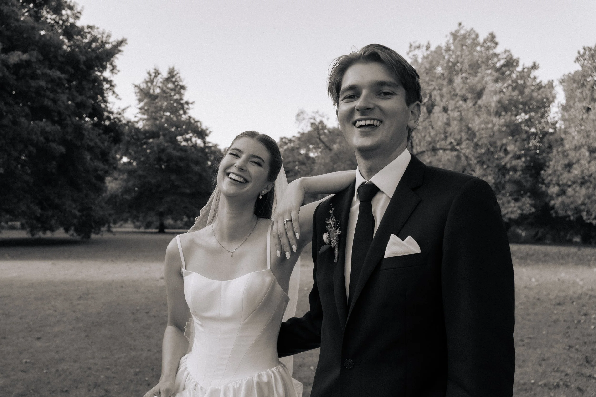 Black-and-white photo of a smiling bride and groom outdoors, with trees in the background. The bride wears a white dress with thin straps and a necklace, and the groom wears a black tuxedo with a tie and pocket square.