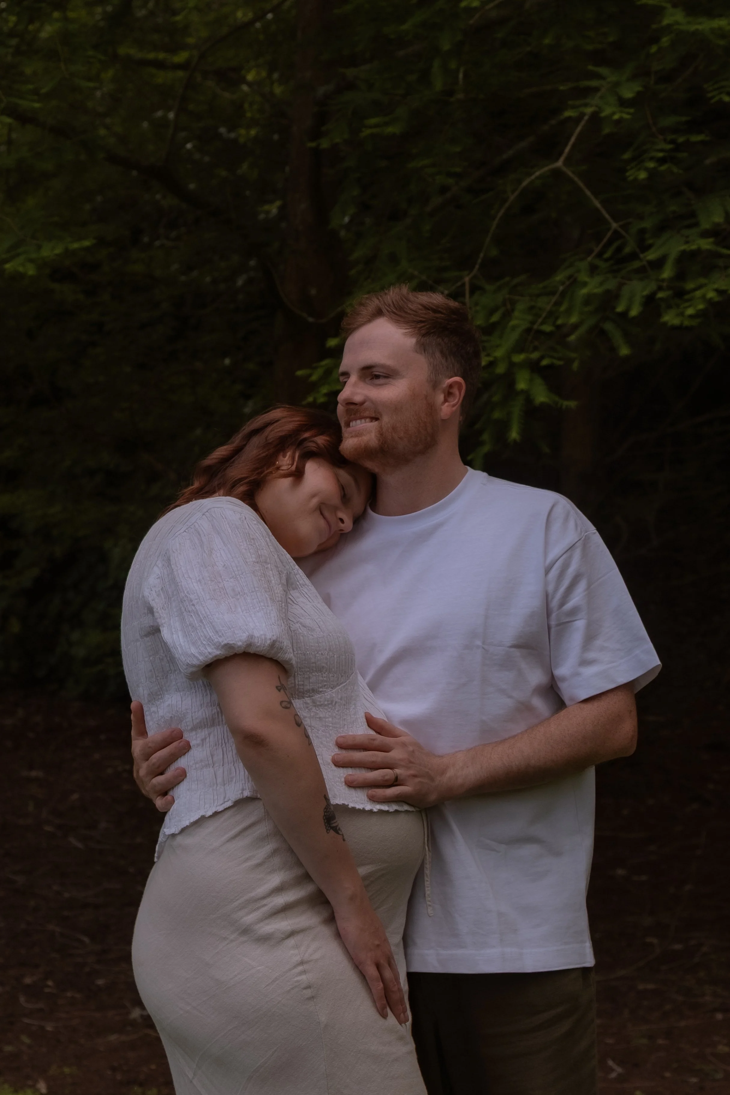 A pregnant woman and a man standing close together outdoors in a wooded area, with the woman resting her head on the man's shoulder and both smiling softly. Maternity Photography. Hamilton New Zealand Photographer