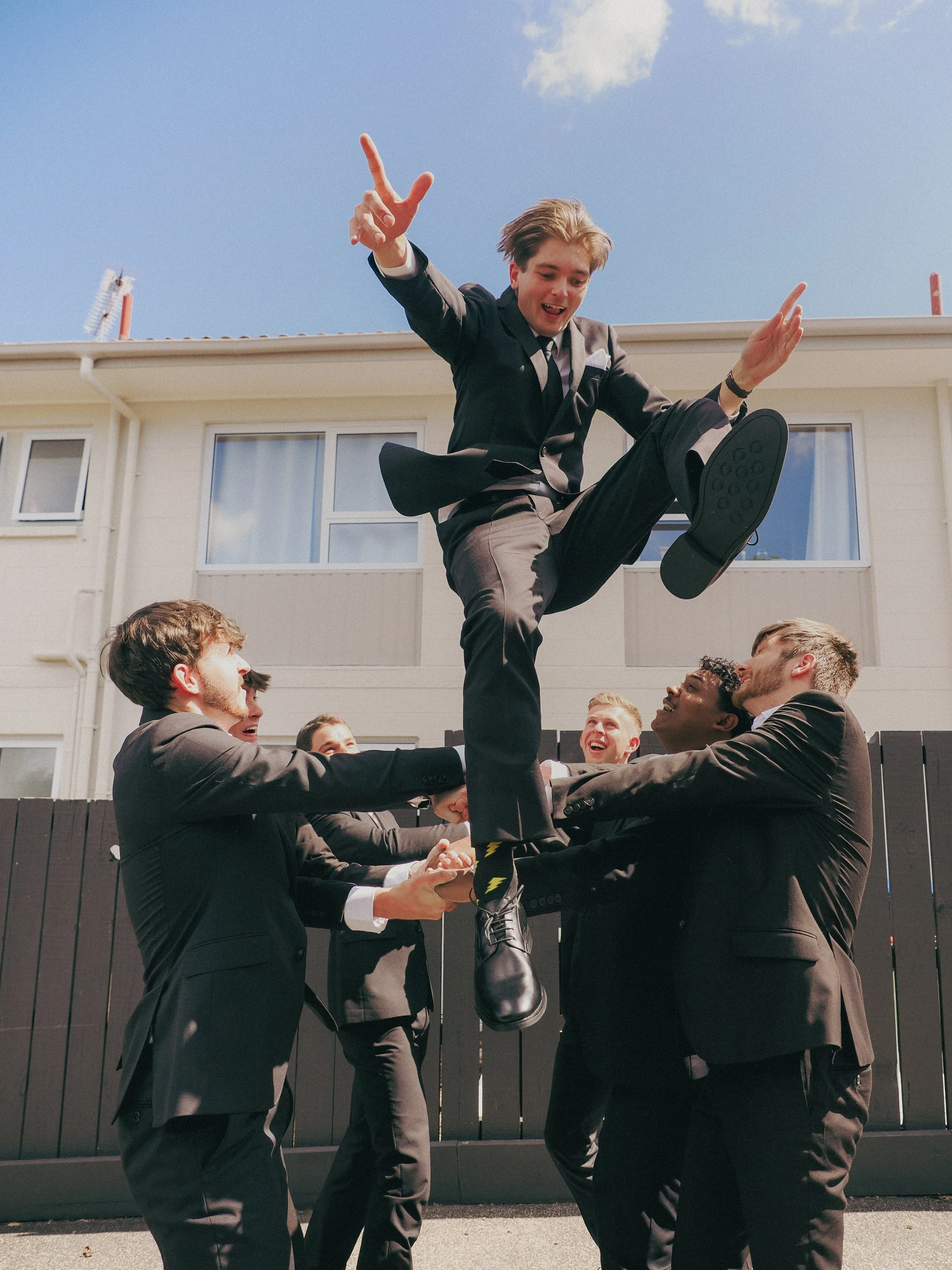 Six men in suits with one in a tie are holding up a young man in a suit with a tie and colorful socks in front of a white building, with a clear sky overhead.