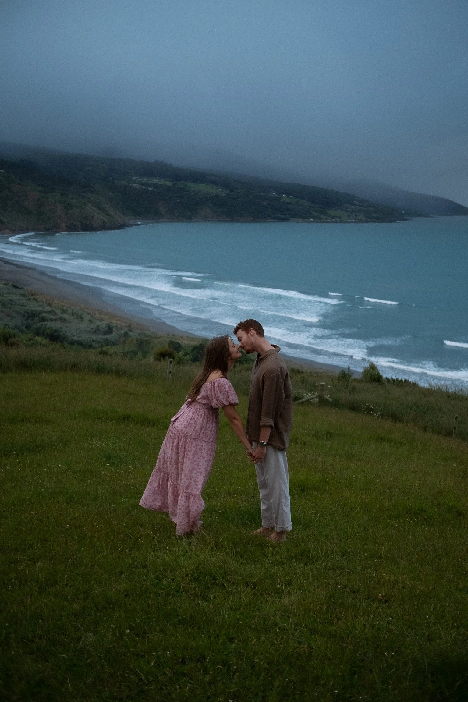 Couple holding hands on a grassy hill with a beach and ocean in the background under a cloudy sky. Couples photography raglan New Zealand. Engagement photographer Hamilton New Zealand. Whangarei New Zealand Photographer 