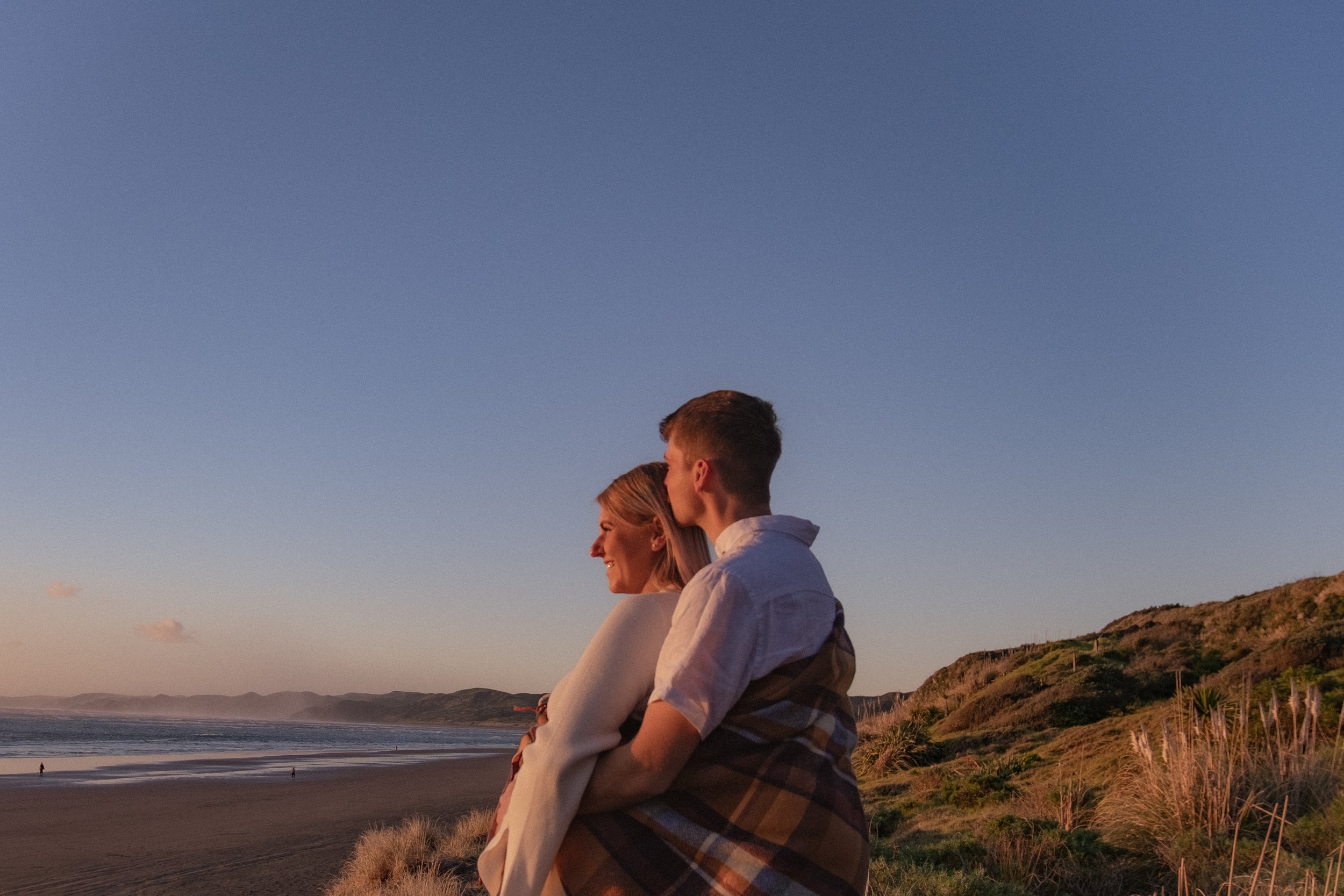 A young couple sitting on a beach at sunset, with the man kissing the woman on the forehead, both smiling and enjoying the view of the ocean and mountains. Maternity Shoot. Raglan New Zealand Photographer
