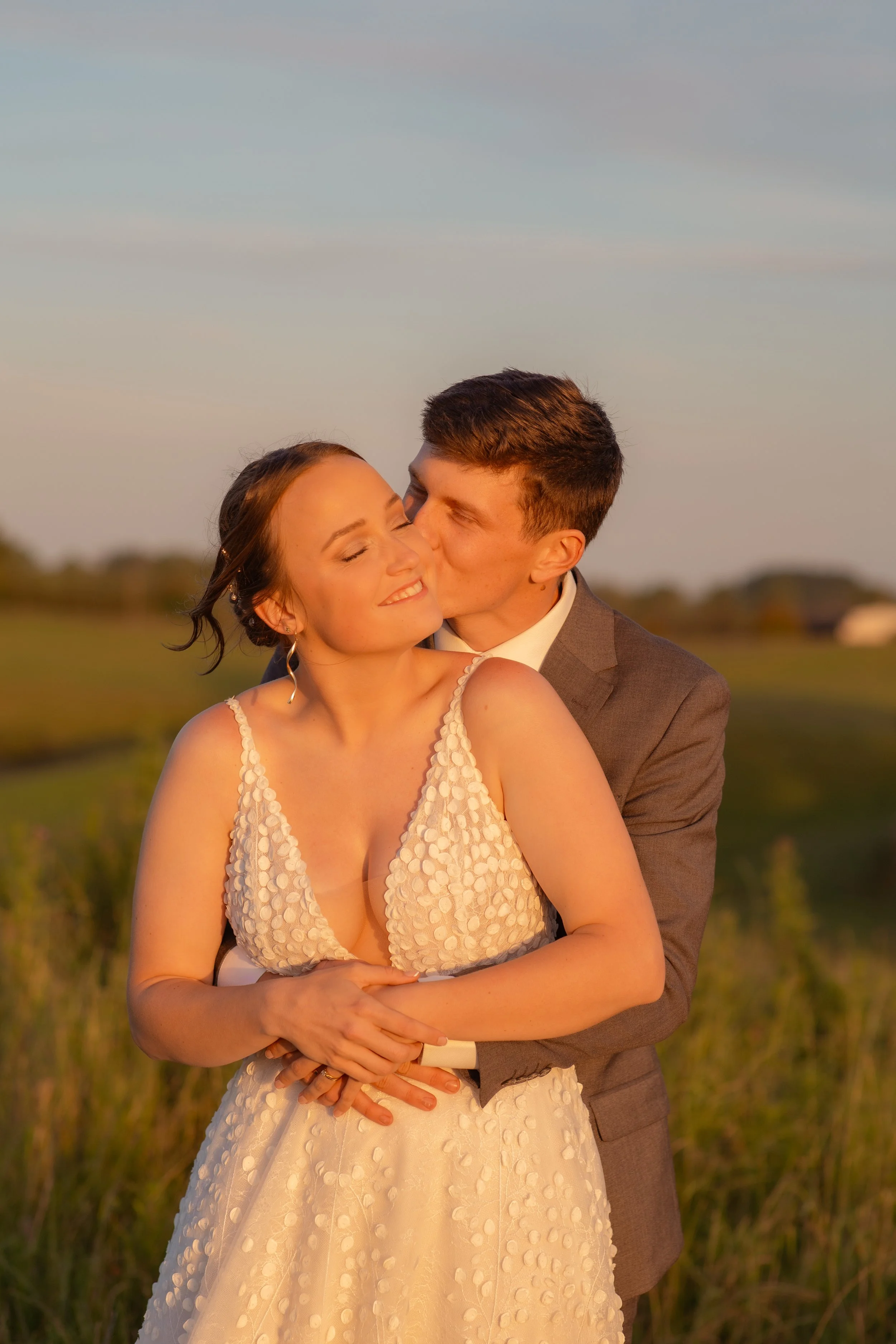 A couple dressed in formal attire sharing a romantic moment outdoors during sunset, with the man kissing the woman on her cheek.  Wedding Photography Waikato New Zealand. Wedding Photography Hamilton New Zealand. Northland Wedding Photography