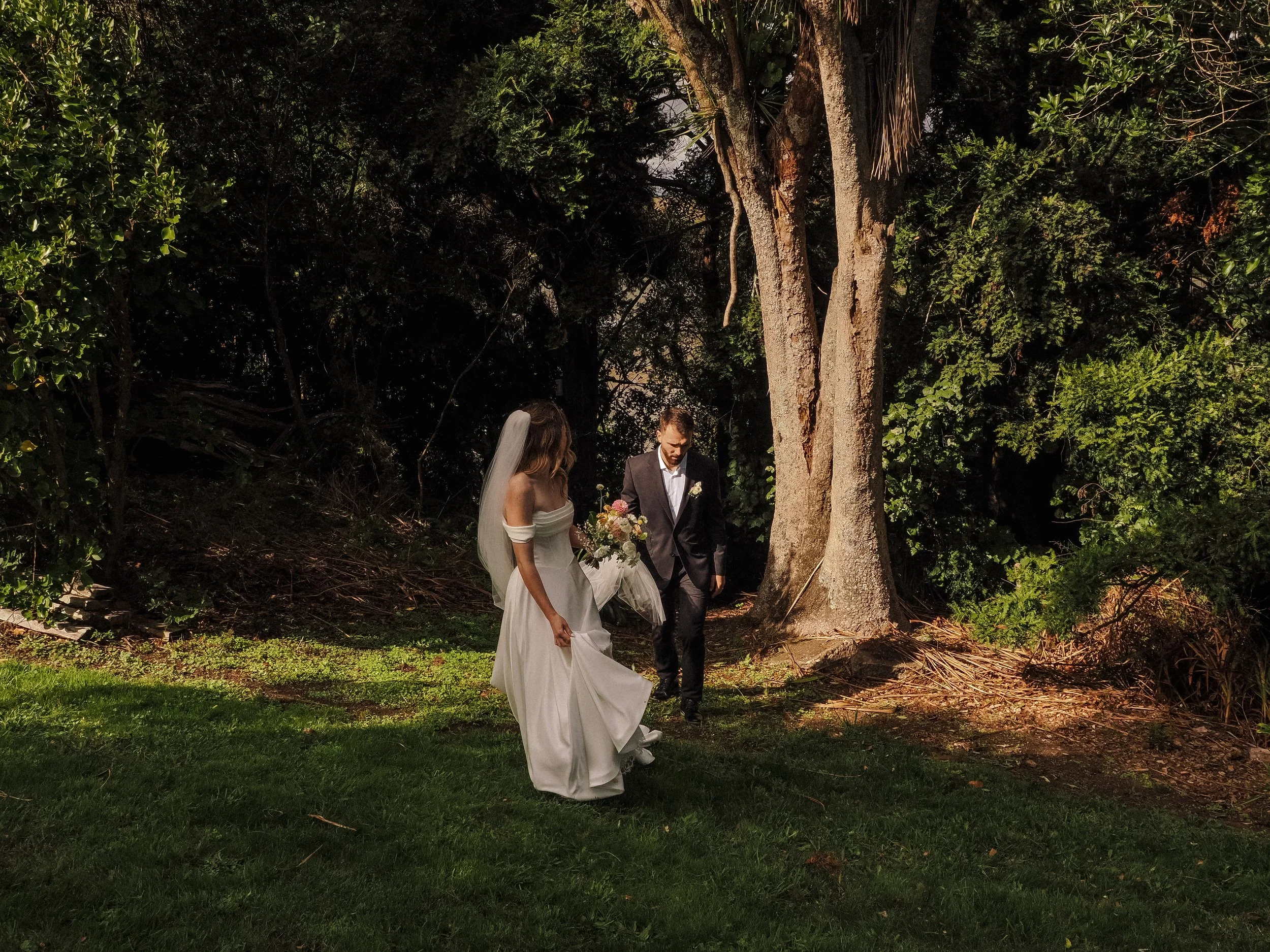 A bride in a white wedding dress and veil holding a bouquet walks beside a groom in a dark suit outside in a wooded area.  Wedding Photography Waikato New Zealand. Wedding Photography Hamilton New Zealand. Northland Wedding Photography