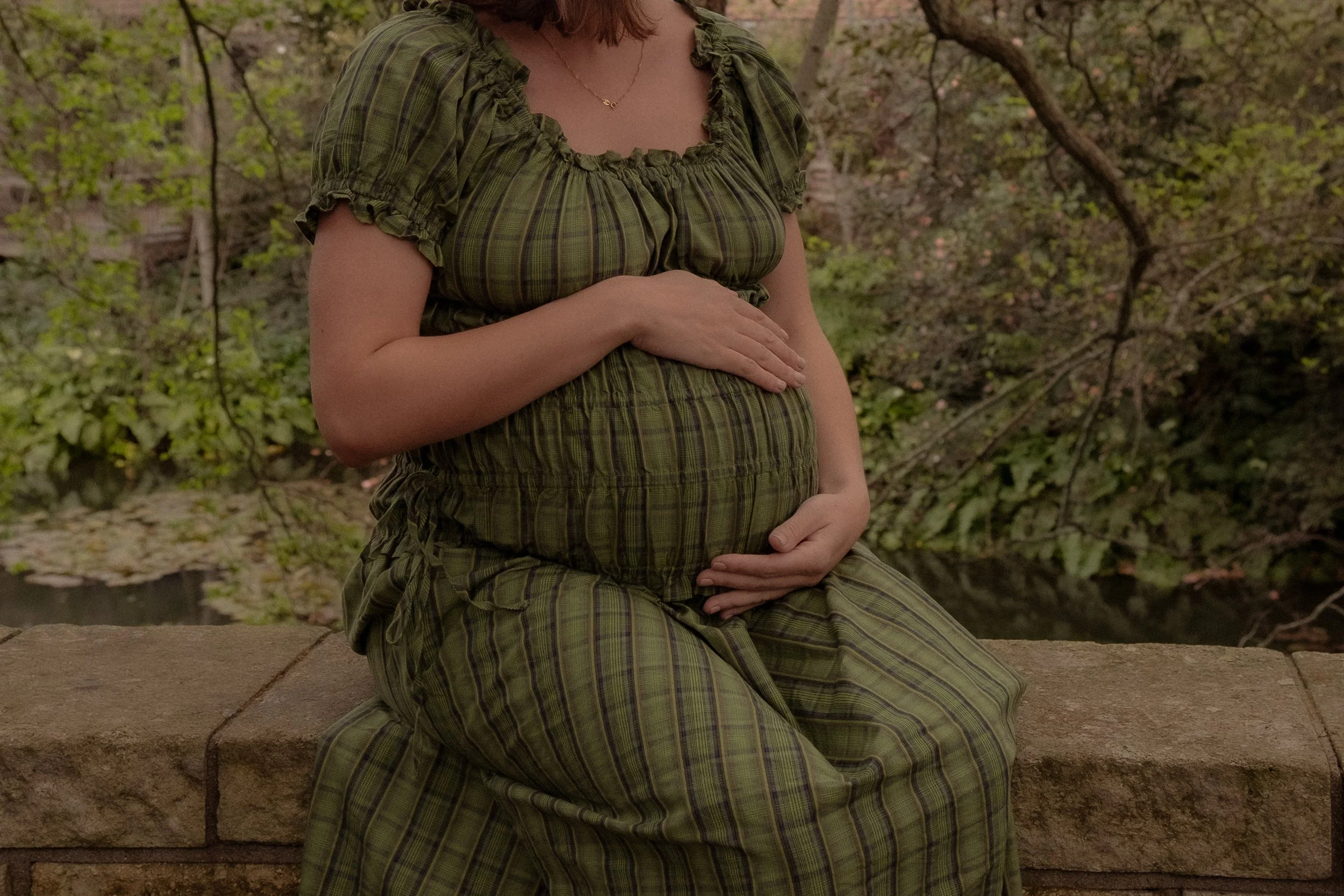 A woman sitting on a stone ledge outdoors, holding her pregnant belly with both hands, wearing a green plaid dress with short puffed sleeves, surrounded by trees and greenery. Maternity photography Hamilton New Zealand