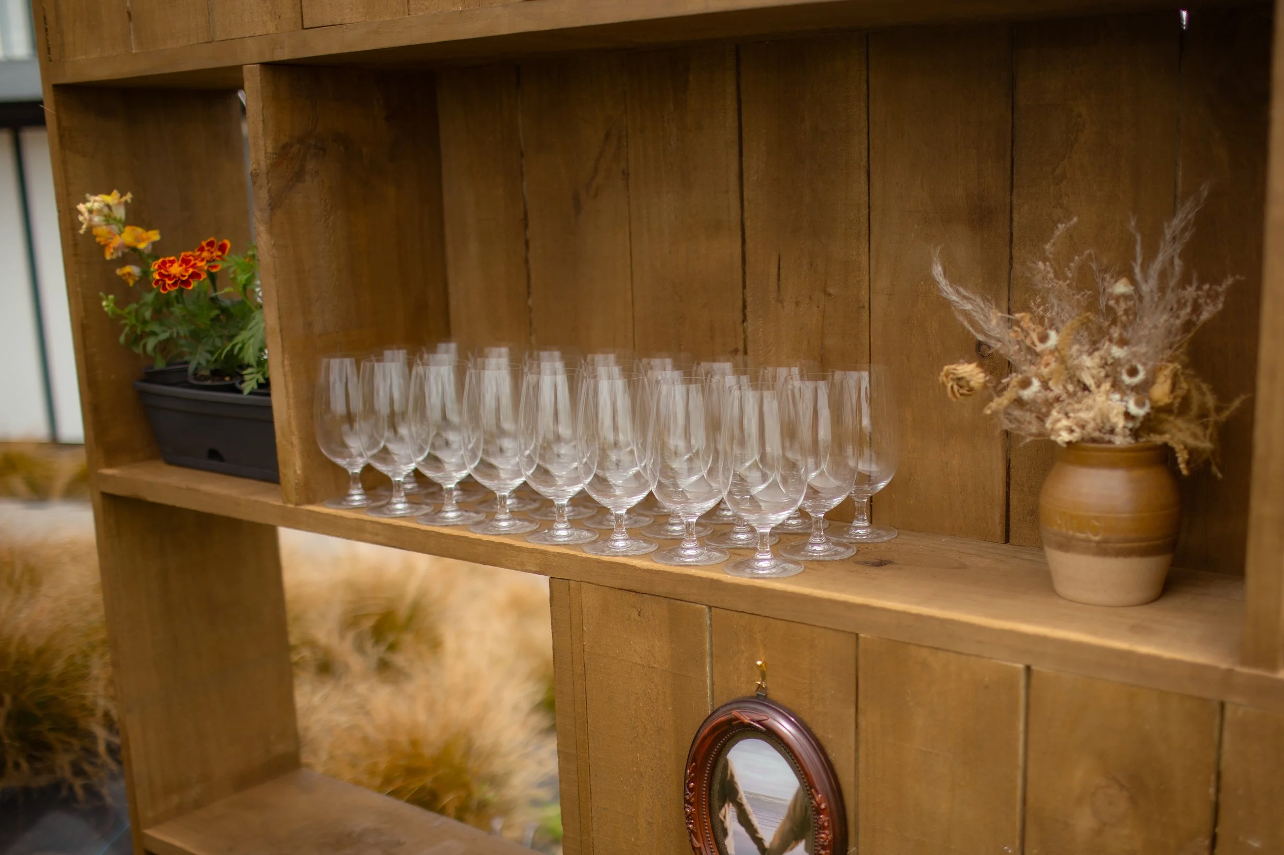 A wooden shelf holding multiple empty wine glasses, a small pot with dried flowers, and a plastic container with colorful flowers, with a blurred animal or fur in the background.  Wedding Photography Waikato New Zealand. Wedding Photography Hamilton