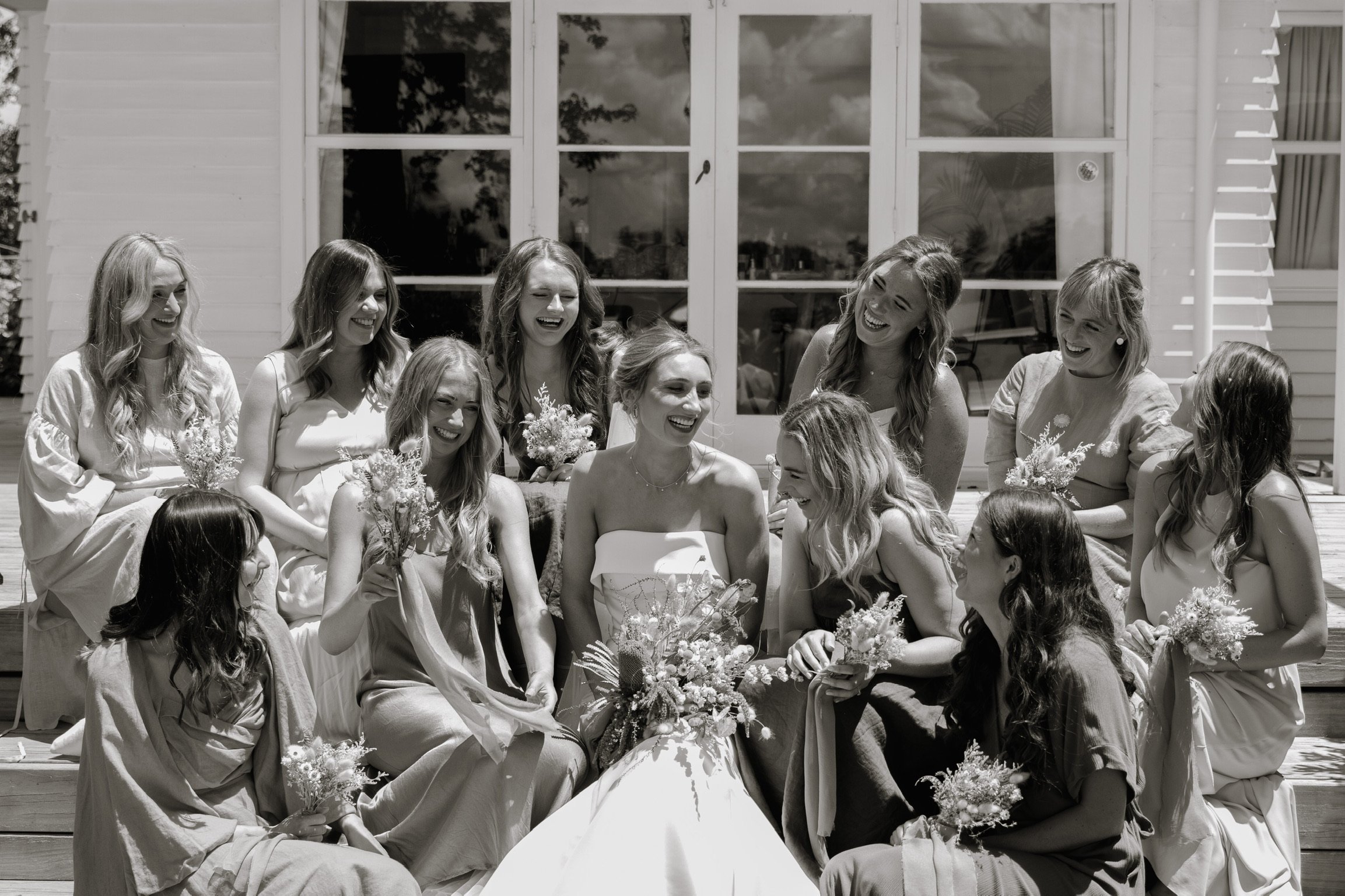 Black and white photo of fifteen women, possibly a bride and her bridesmaids, sitting on steps outside a building with large windows, laughing and holding bouquets of flowers.  Wedding Photography Waikato New Zealand. Wedding Photography Hamilton 