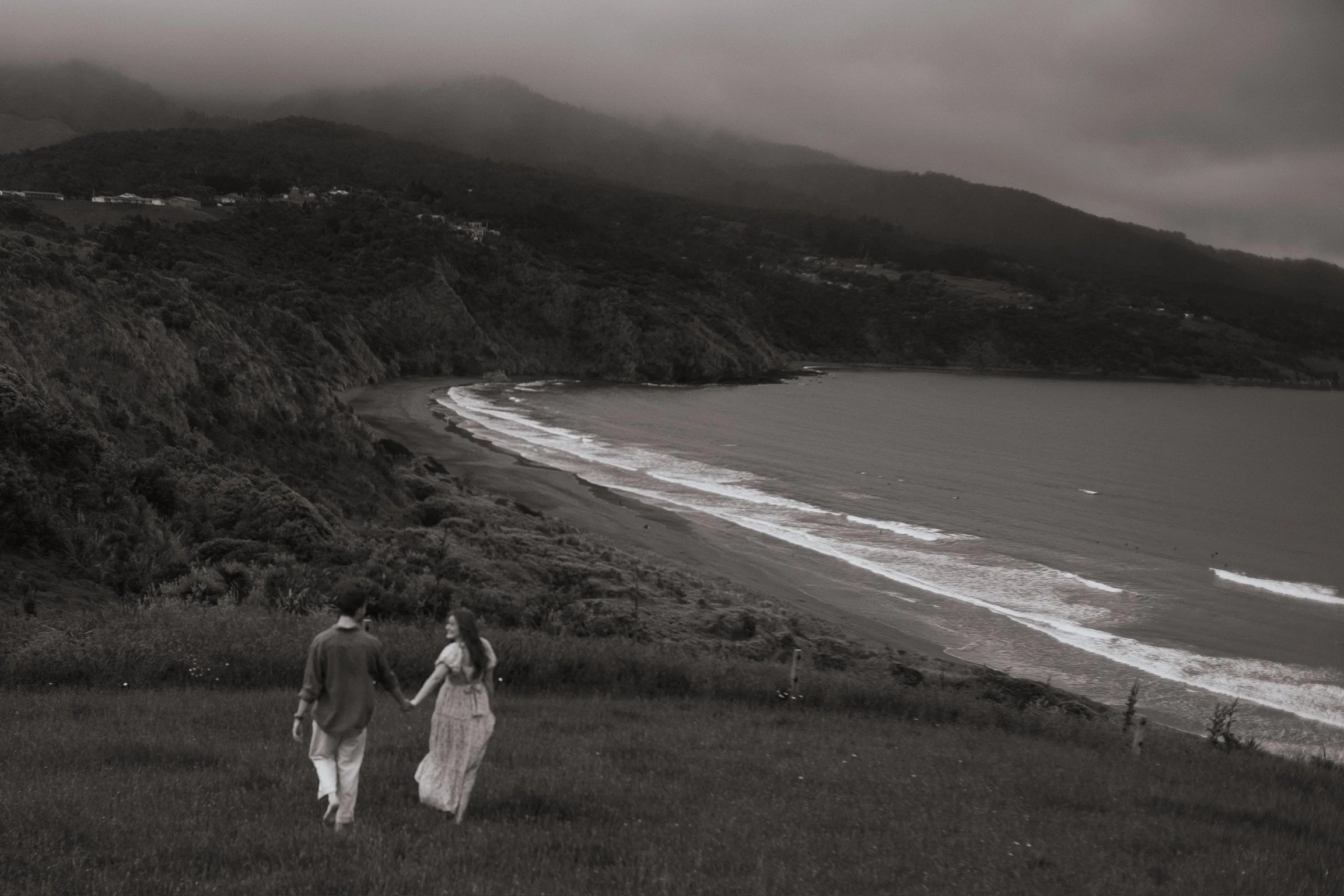 A couple holding hands walking on a grassy hill overlooking a beach with waves, cliffs, and mountains in the background under cloudy sky. Couples photography Raglan New Zealand. Engagement photography Hamilton New Zealand. Whangarei Photographer