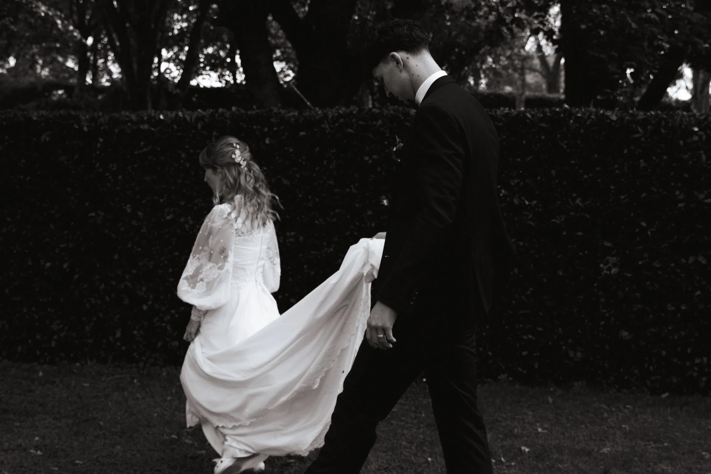 A black-and-white photo of a bride and groom outdoors. The bride, wearing a lace wedding dress, is walking with her head down. The groom, dressed in a tuxedo, is holding her wedding train as they walk past a dark hedge and trees in the background.