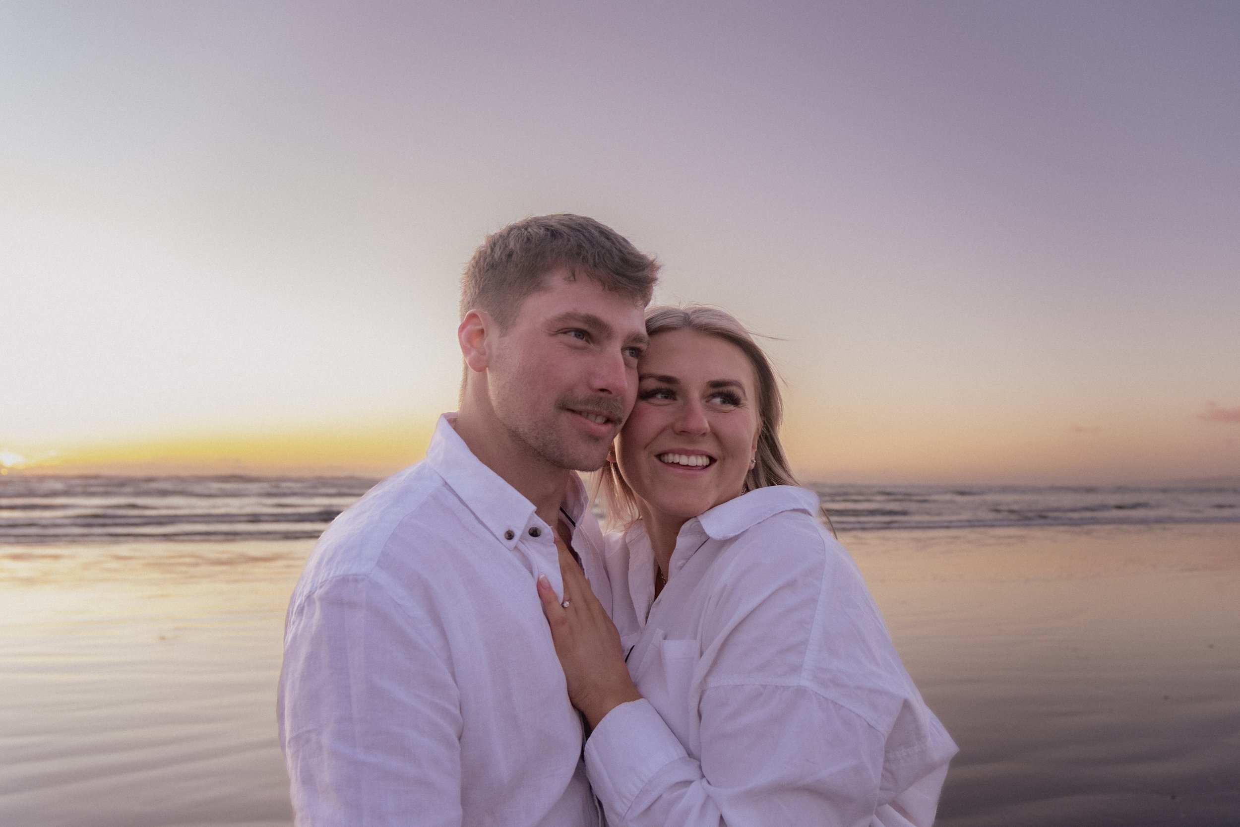A happy couple hugging on a beach during sunset, smiling and looking towards the horizon. Maternity Photography. Raglan New Zealand Photographer