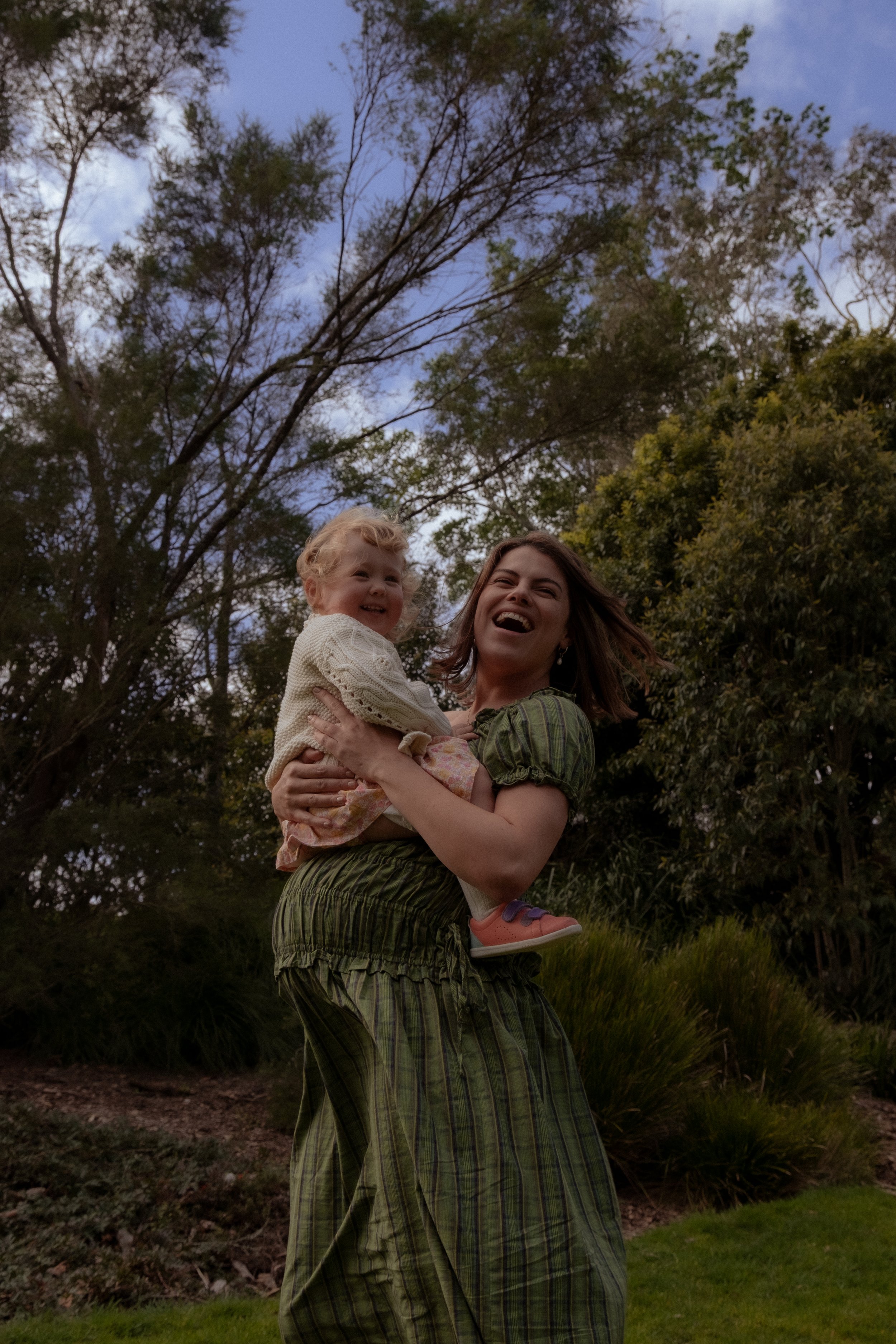A woman is holding a smiling young girl in her arms outdoors, surrounded by trees and bushes, during daylight hours. Family photographer, Maternity photography, Hamilton New Zealand Photographer