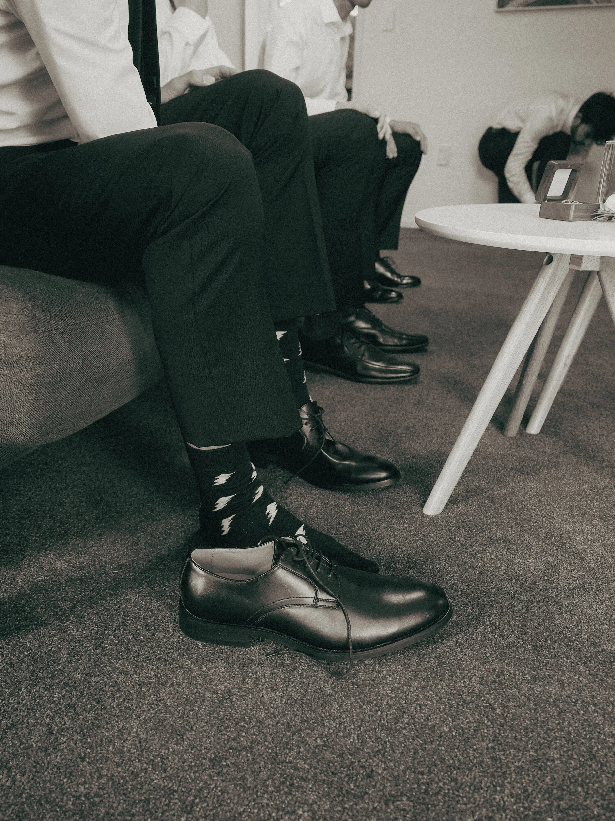 Men sitting on a couch, wearing black dress shoes, black pants, and white socks with black lightning bolt patterns, with one shoe removed and placed on the carpet. There is a white side table with a tissue box and other items on it.
