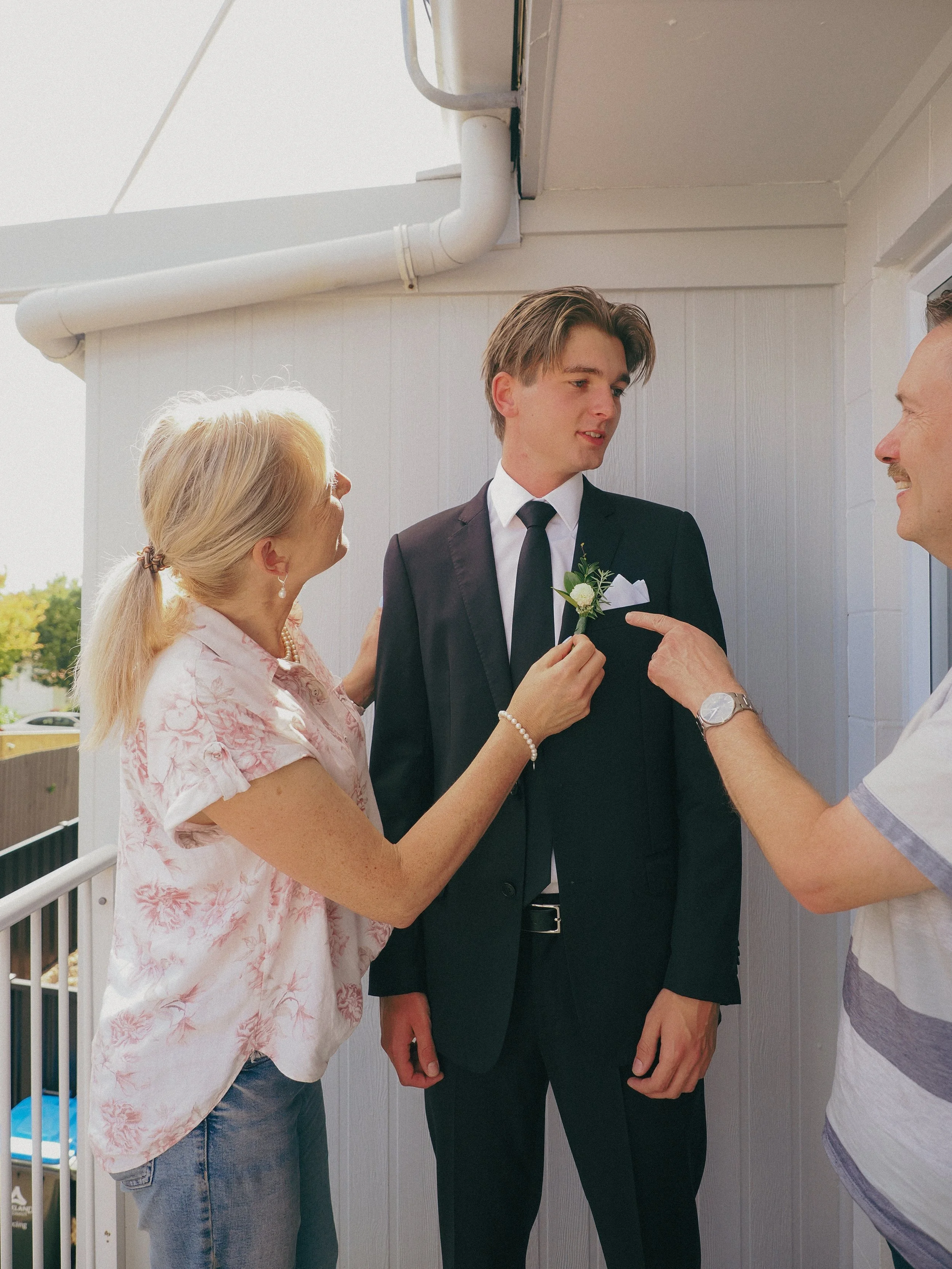 A young man in a black suit and tie is standing outside a white building, receiving a boutonniere from a woman with blonde hair wearing a pink patterned blouse. An older man with a watch is pointing at his boutonniere, and another man in a striped sh