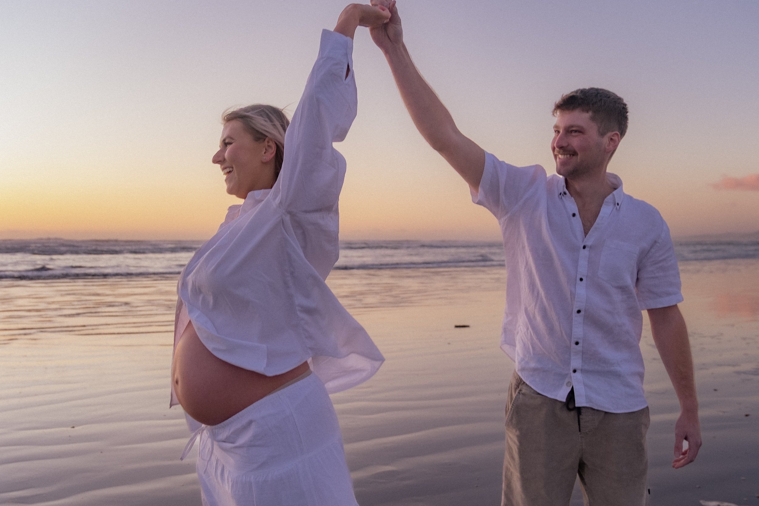 A pregnant woman and a man dancing and holding hands on a beach during sunset, smiling and enjoying each other's company. Maternity Photography. Raglan New Zealand Photographer