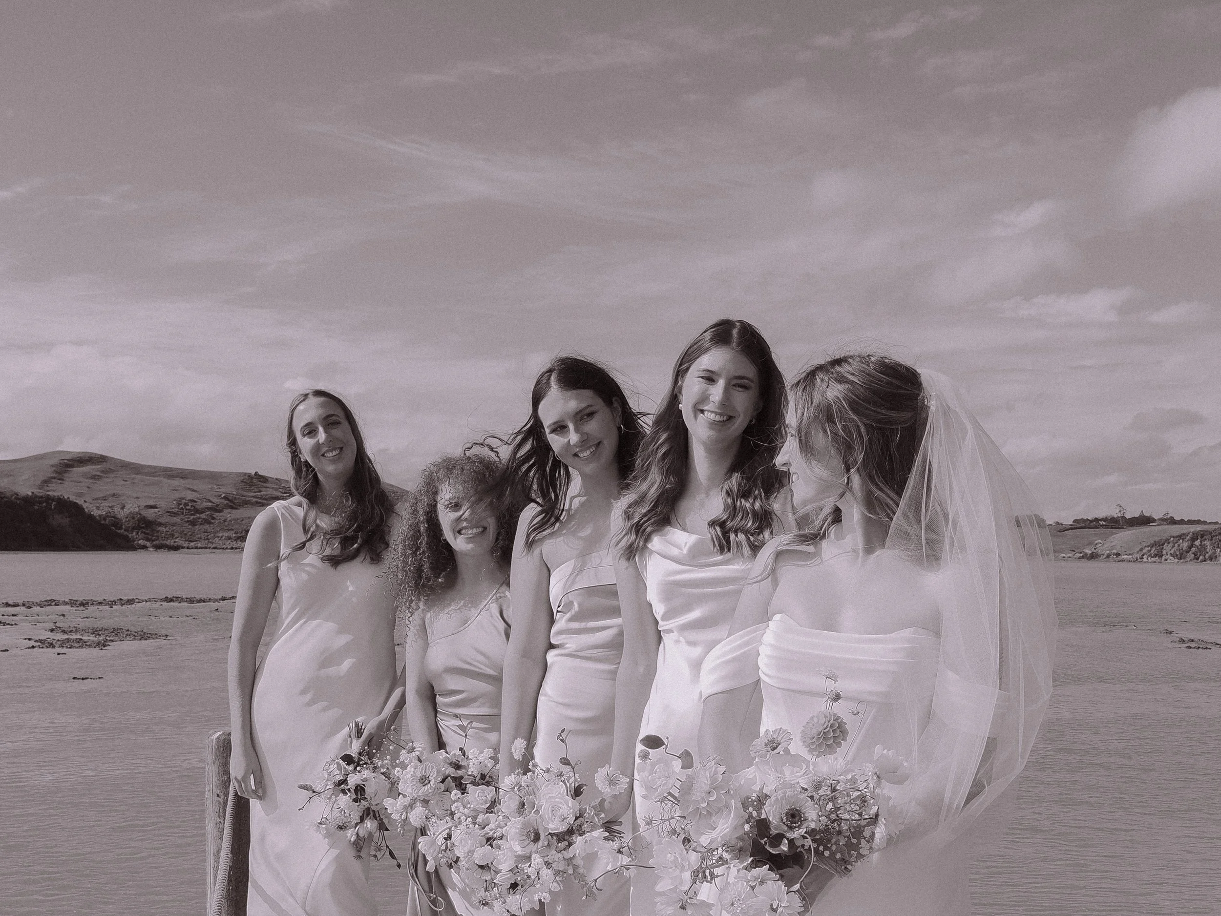 A group of five women on a beach, with one woman in a wedding dress and veil, holding a bouquet, and four women in light-colored dresses, smiling and standing in a row.  Wedding Photography Waikato New Zealand. Wedding Photography Hamilton 