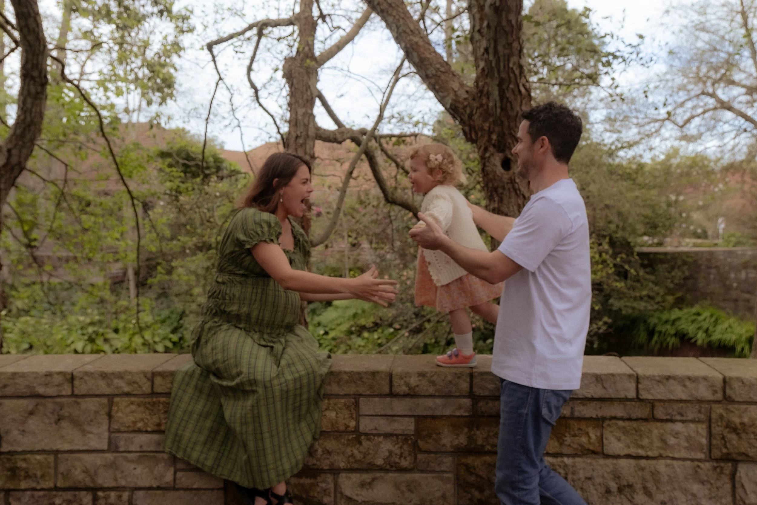 Family enjoying outdoor time, girl and woman reaching for each other, man holding a young girl, in a backyard with trees and a brick wall. Family/Maternity Photography Hamilton New New Zealand