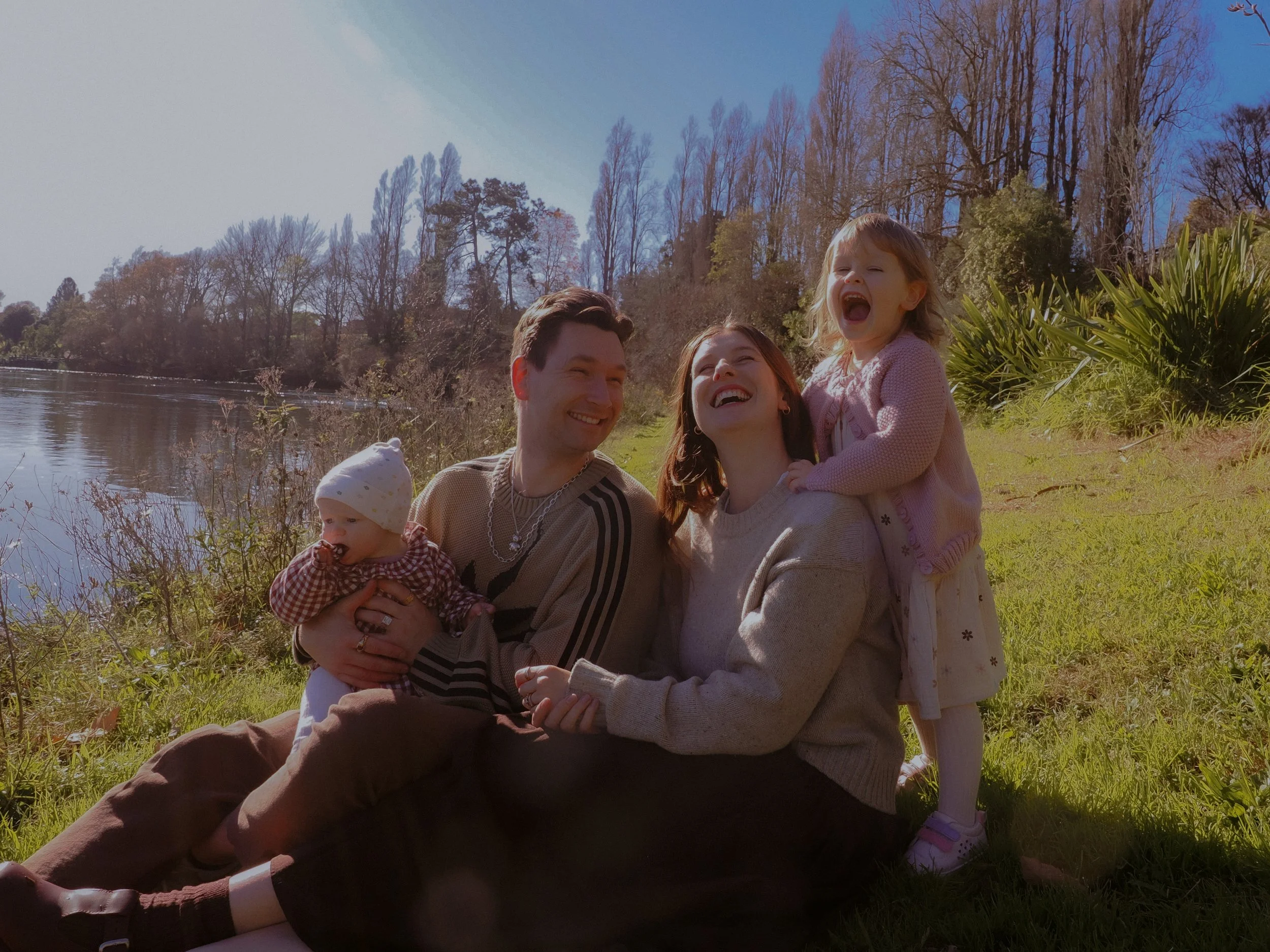 A family of four sitting by a lake, smiling and having fun on a sunny day. The father holds a baby, the mother is sitting next to him, and a young girl is standing behind them, laughing. Family Photography Waikato New Zealand. Family Photography 