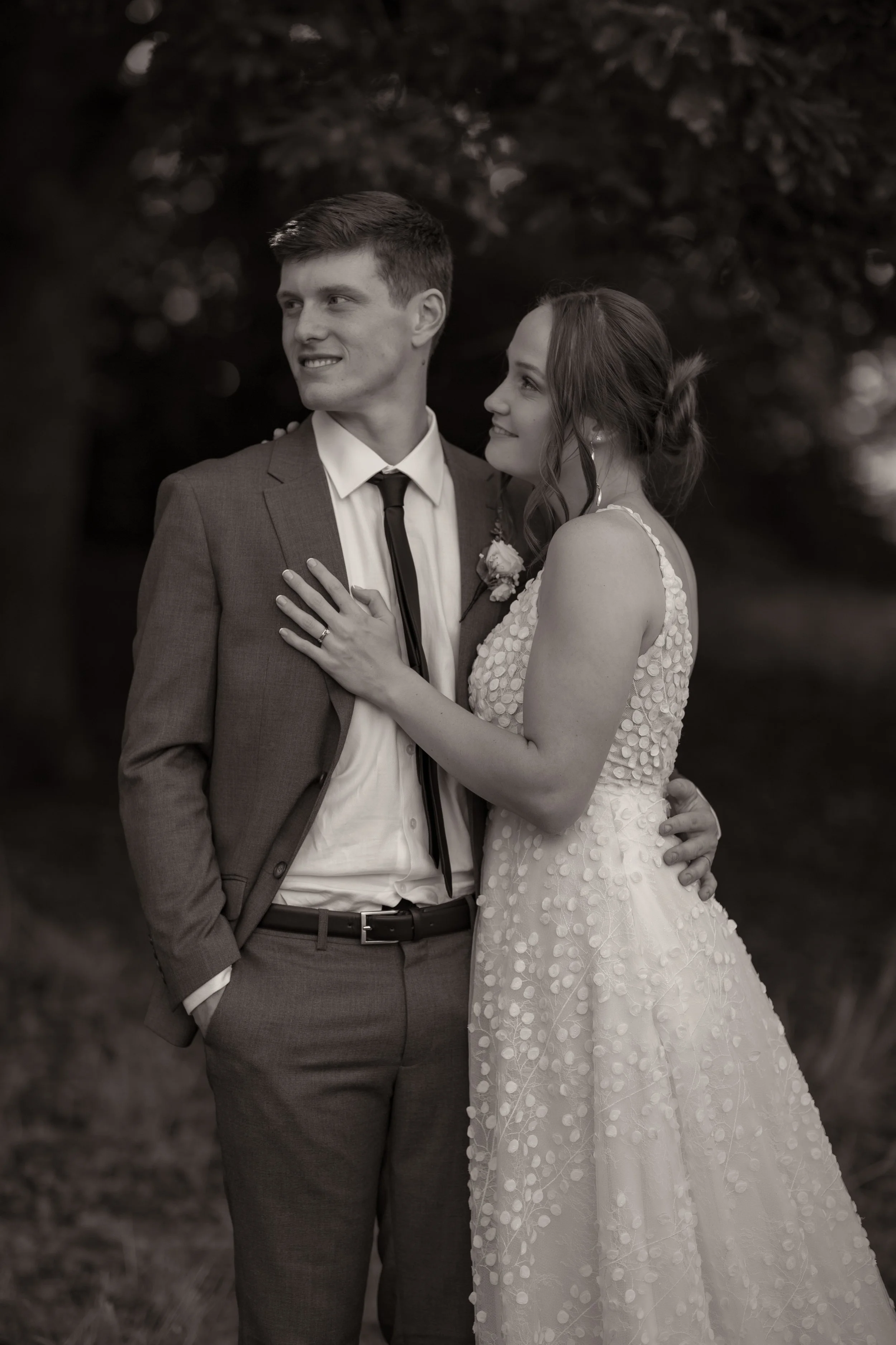 A black and white photo of a newlywed couple standing close together outdoors, with the woman resting her hand on the man's chest, both looking off to the side.  Wedding Photography Waikato New Zealand. Wedding Photography Hamilton New Zealand. North