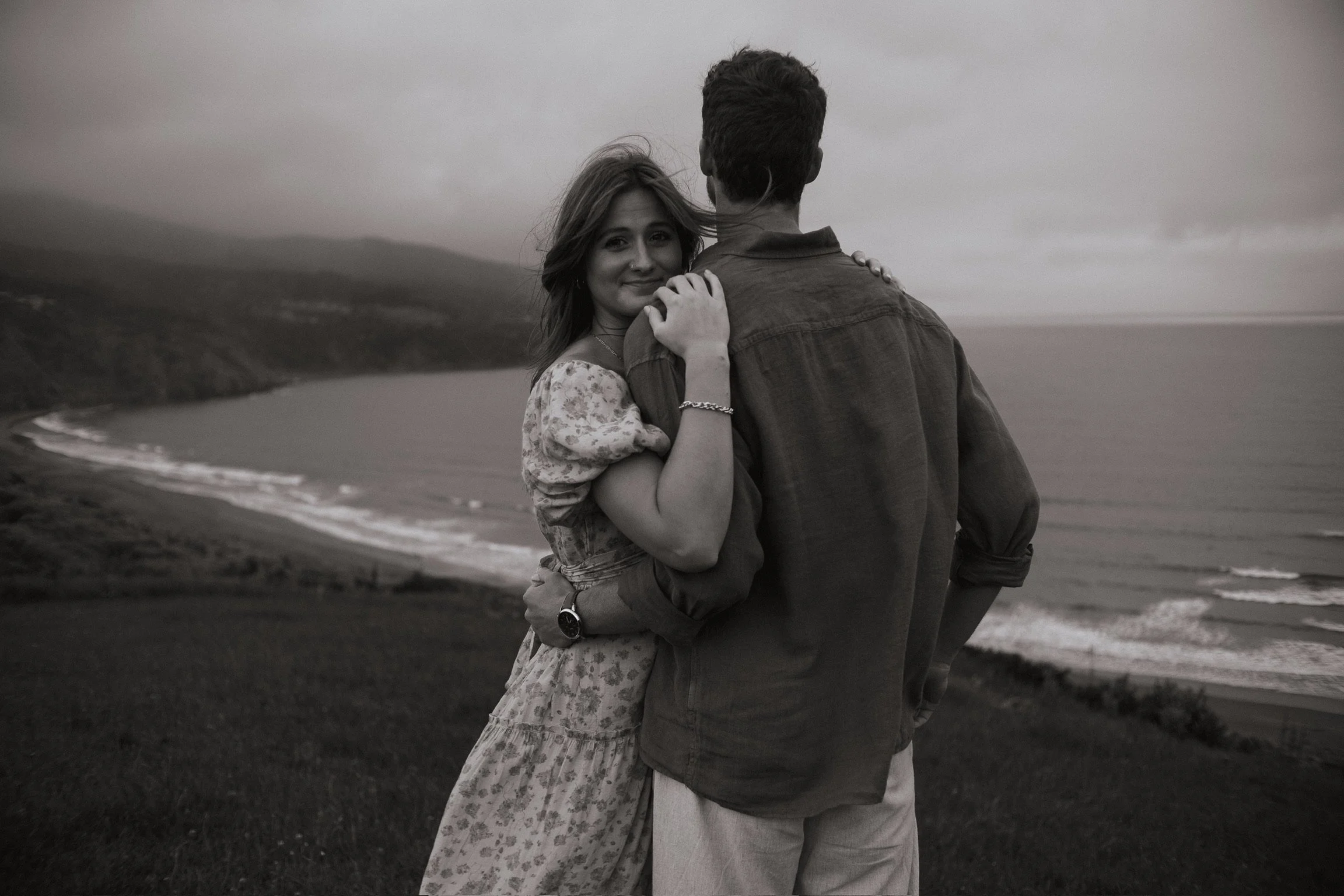 A couple embracing on a grassy hill overlooking the shoreline and ocean at sunset, black and white photograph. Couples photography Raglan New Zealand. Engagement photography Hamilton New Zealand. Whangarei New Zealand Couples photographer