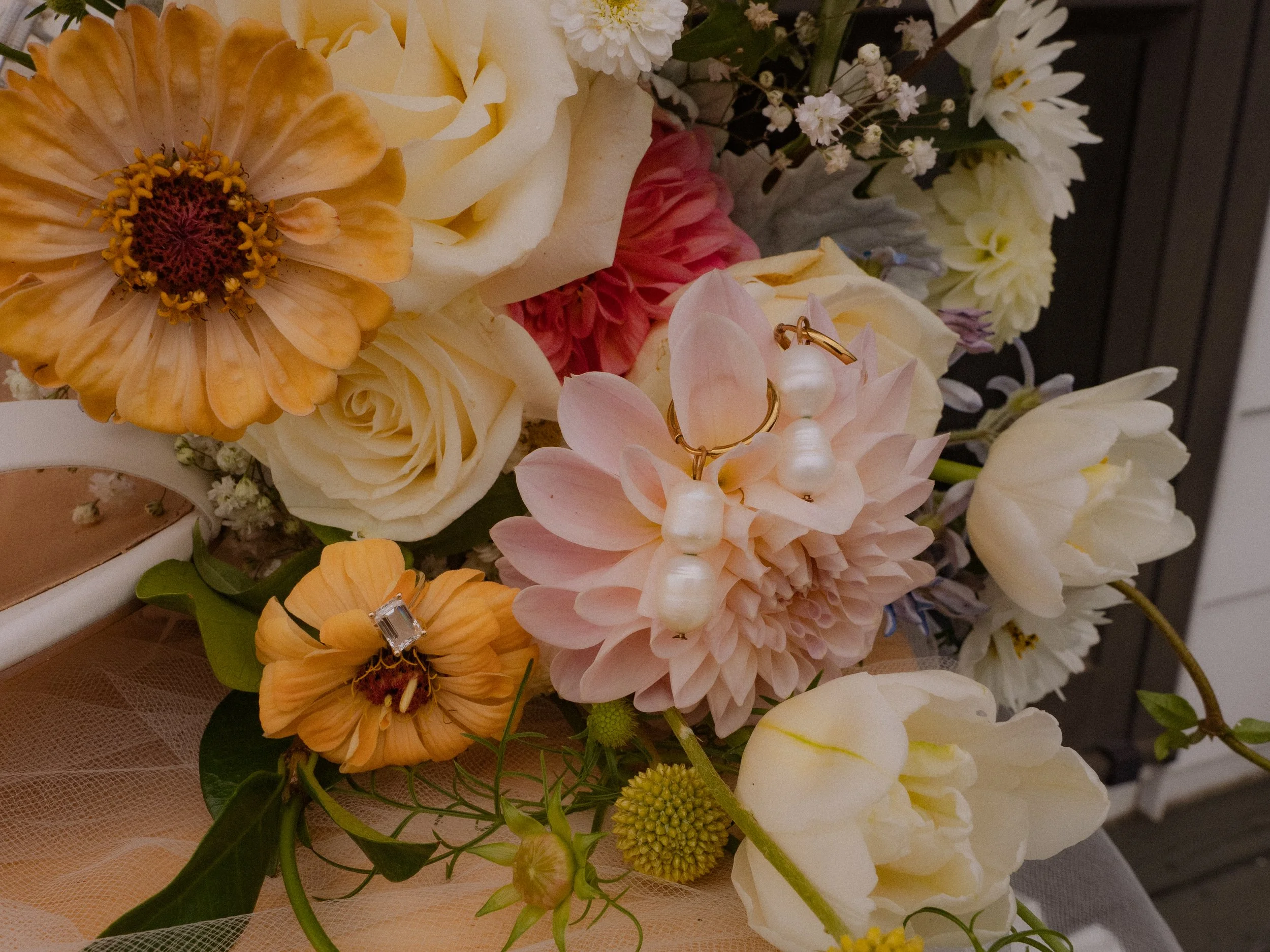A close-up of a floral bouquet with various white, cream, yellow, and peach-colored flowers, including roses, dahlias, and small filler flowers, with a pair of pearl and gold earrings resting on a pink dahlia in the foreground.  Wedding Photography 